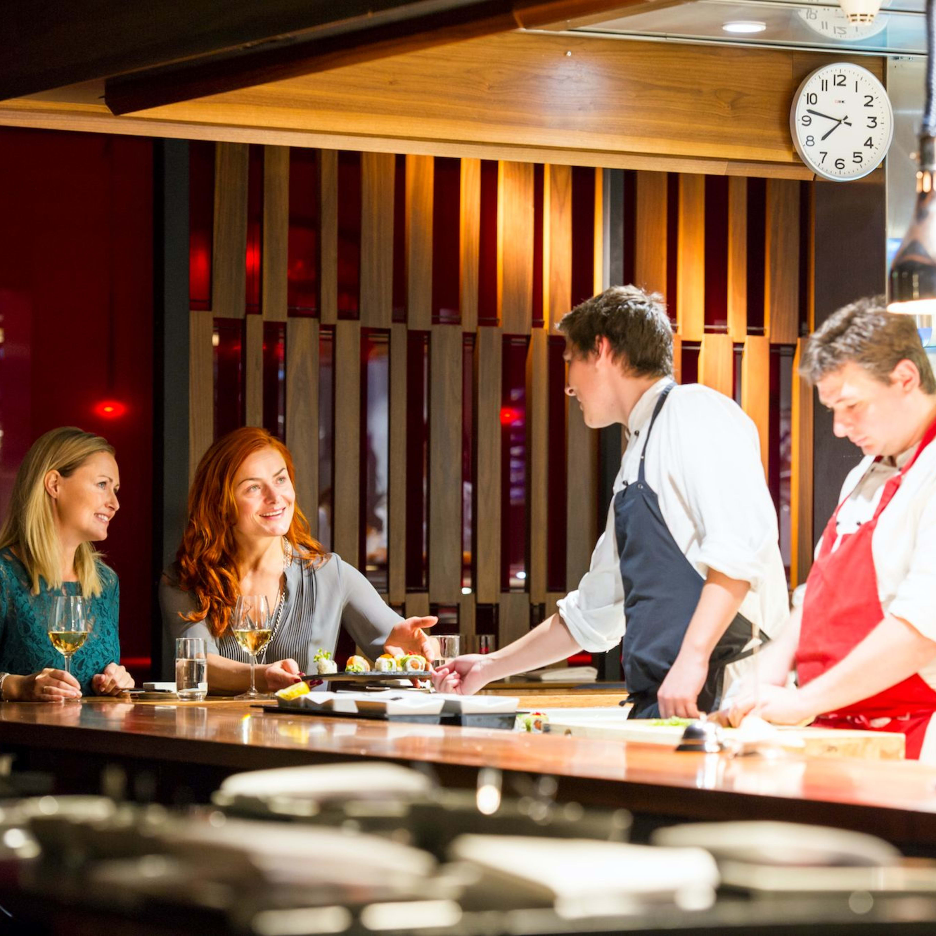 Frauen an der Restaurantbar, die mit dem Koch, der Sushi serviert, in einem modernen, warmen Küchenumfeld interagieren.