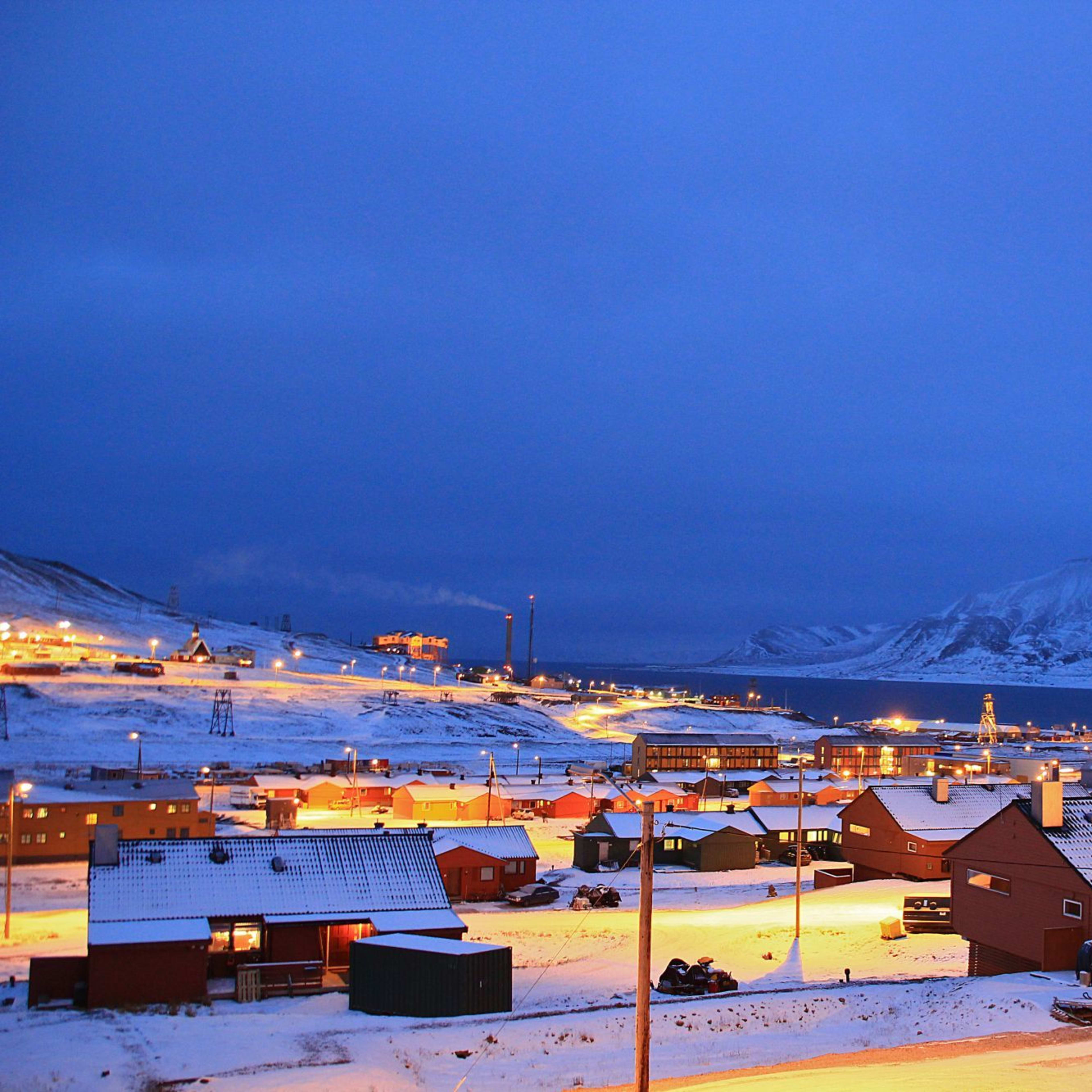 Lite by dekket med snø om natten, varmbelyste hus, gatebelysning som kaster et mykt lys, med snødekte fjell i bakgrunnen.