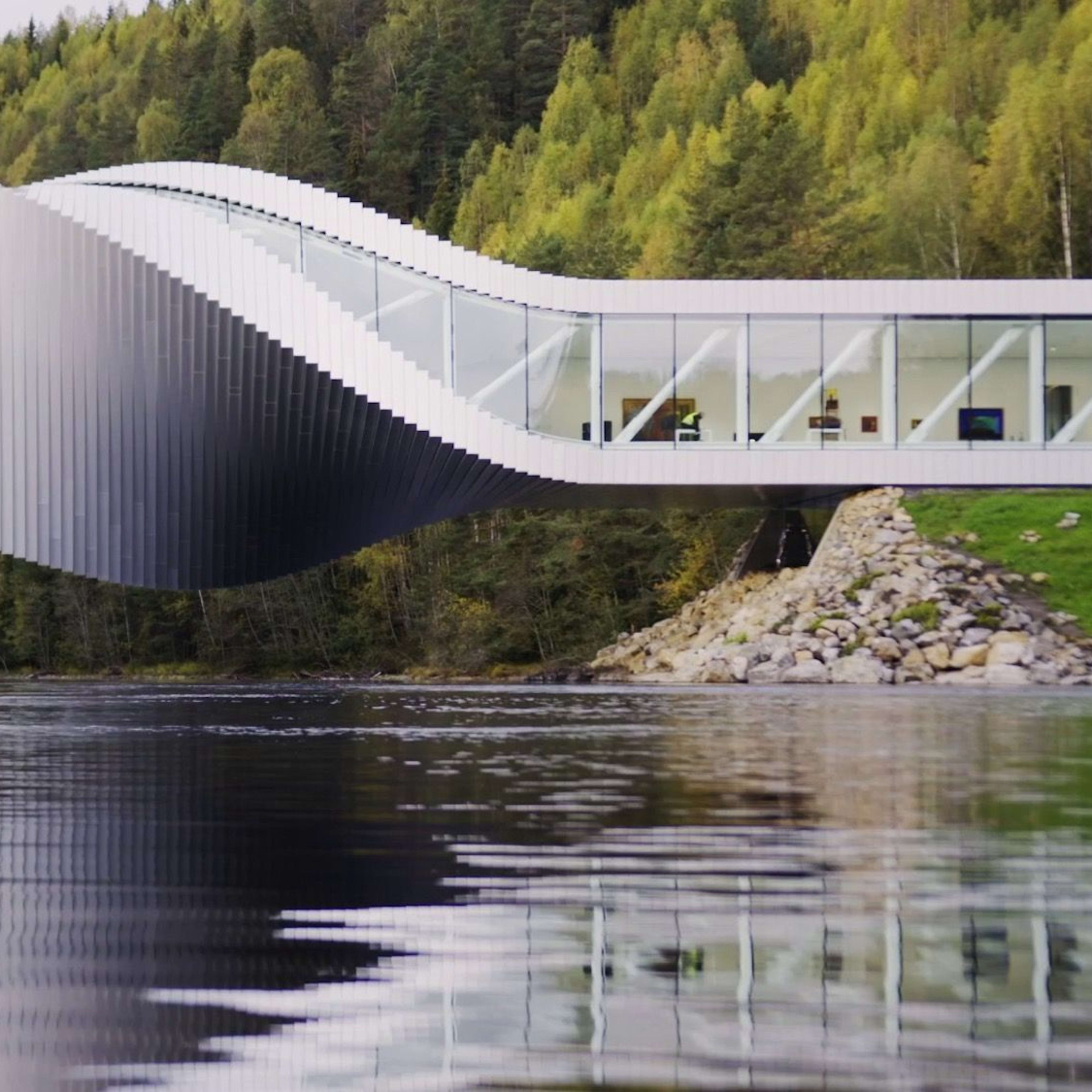 Moderne arkitektonisk struktur over en elv, med bølgelignende design, store glassvinduer og refleksjon i rolig vann.