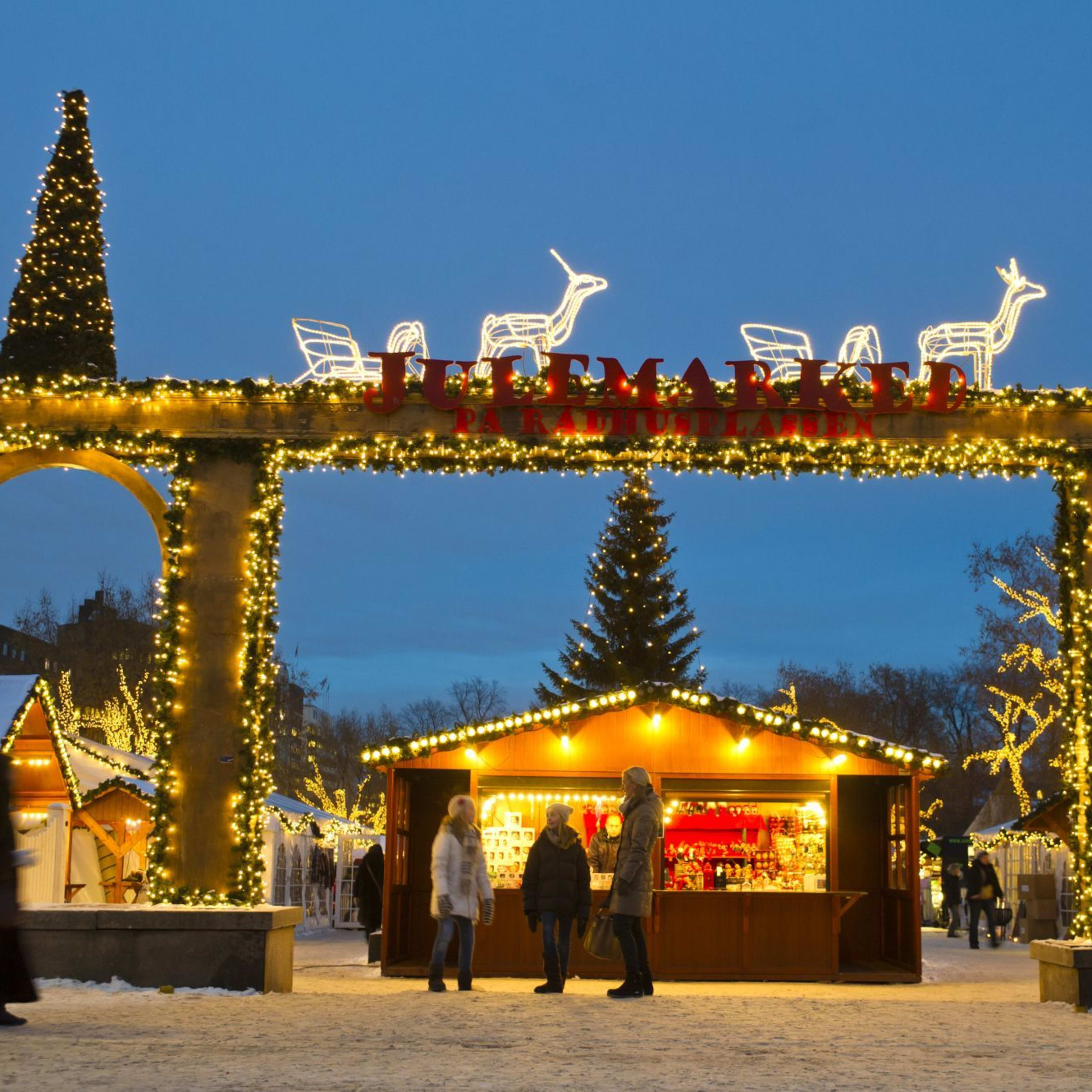 Christmas market entrance with lights and "JULMARKED" sign, reindeer decorations, and festive stalls in snow-covered street.