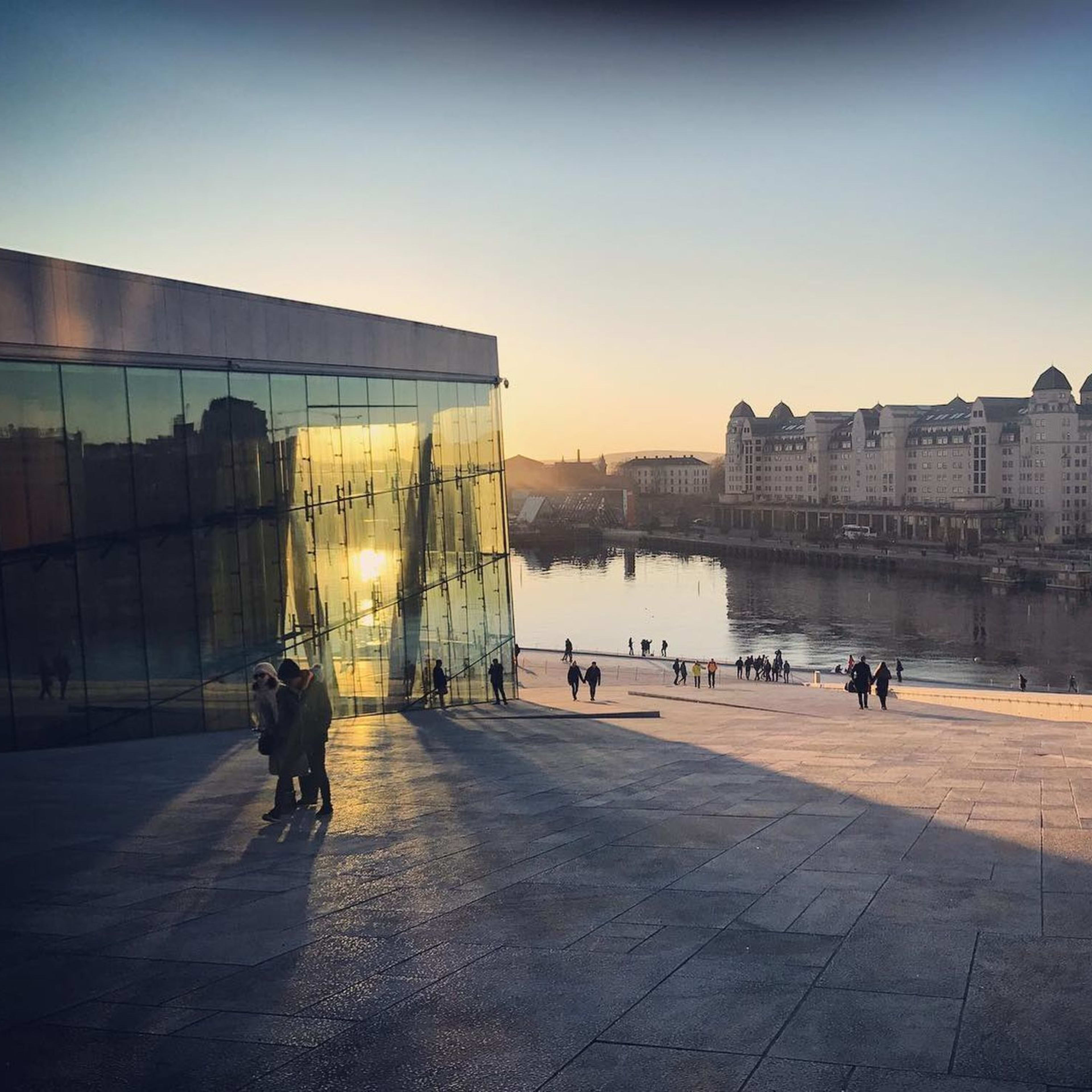 Oslo Opera House with sunset reflecting on glass facade, people walking on marble steps, and city reflections in water.