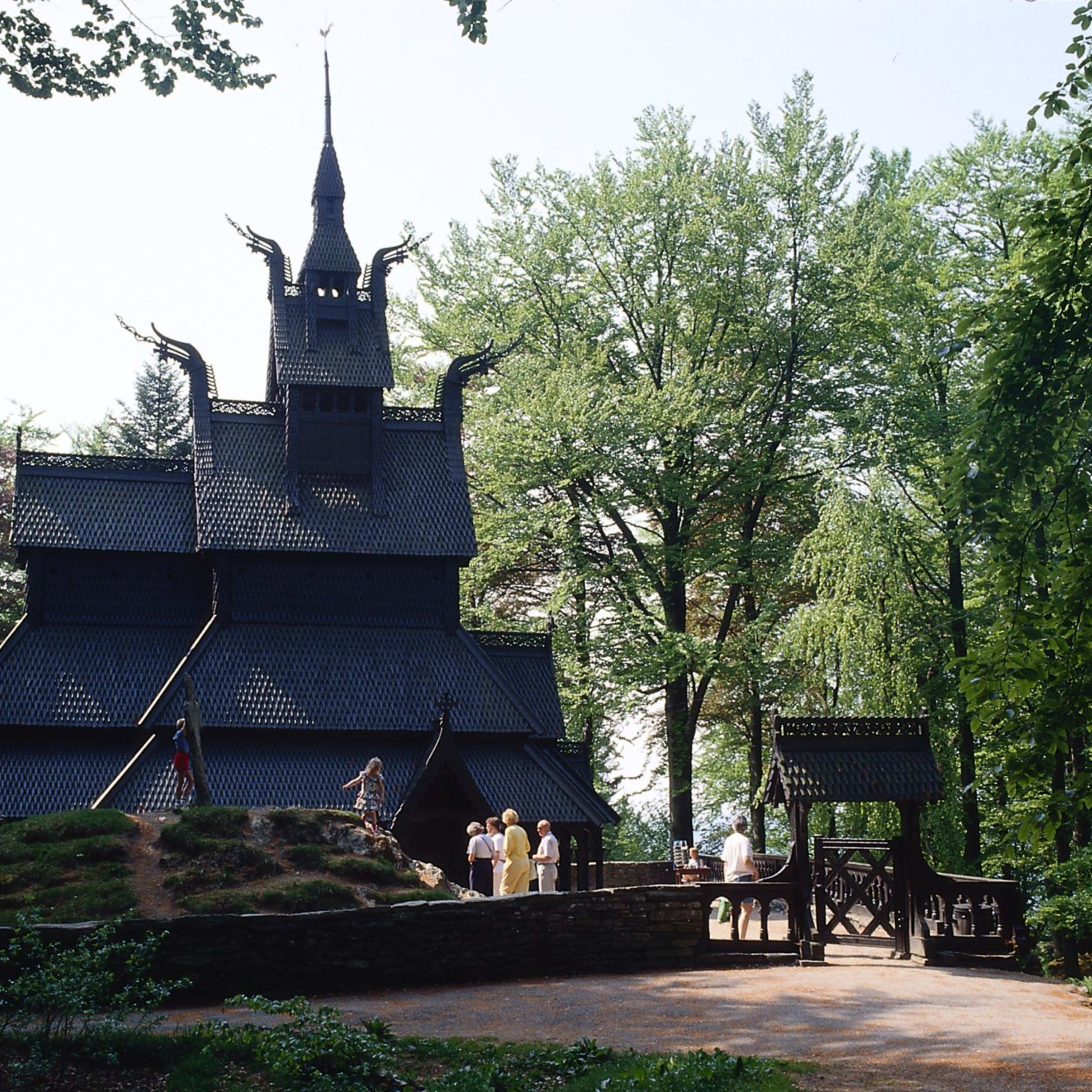 En svart stavkirke omkranset av trær, med intrikate utskjæringer og et flertårnet tak. Barn leker i nærheten.