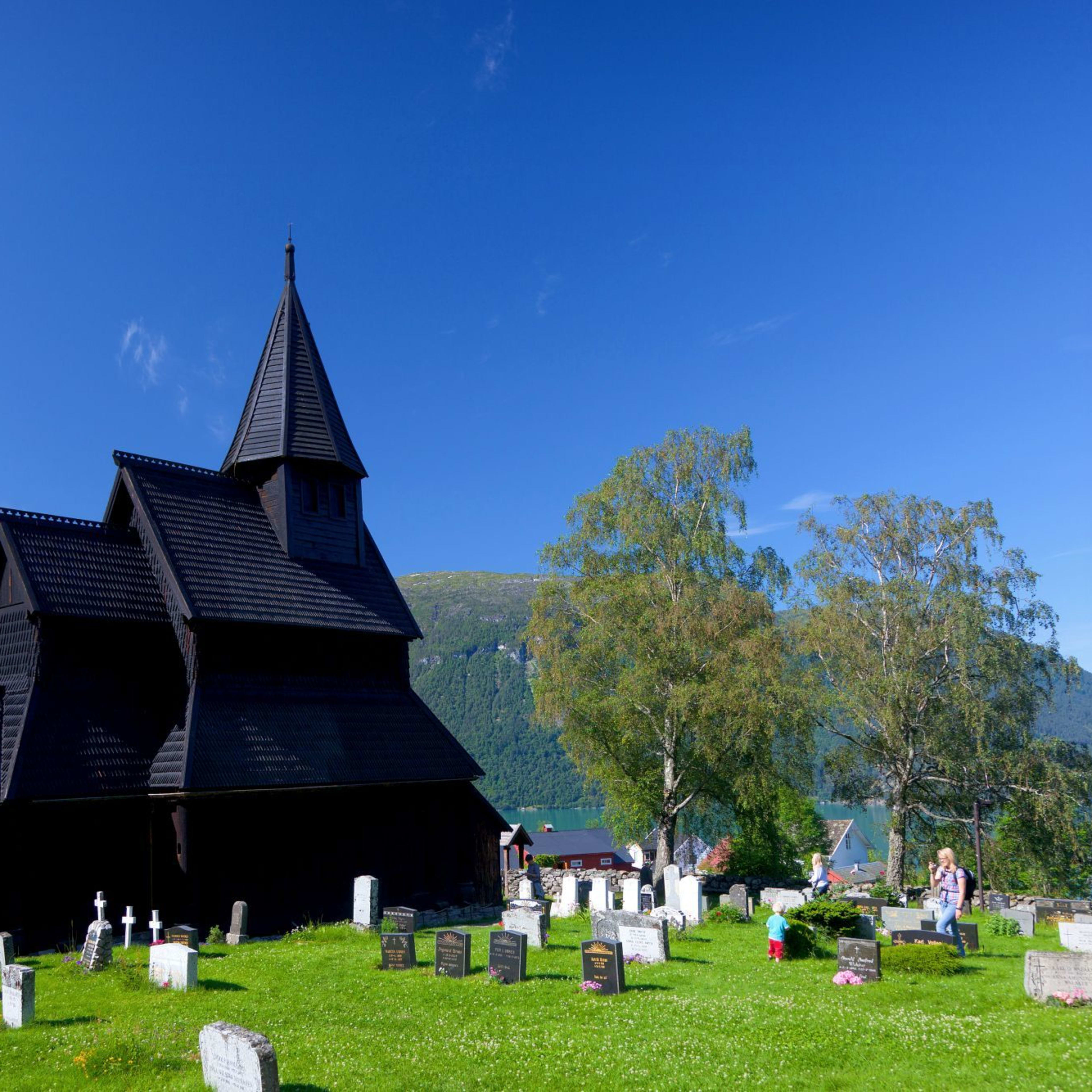En svart trebent stavkirke med bratte tak står mot grønne bakker, en innsjø og en lys blå himmel.