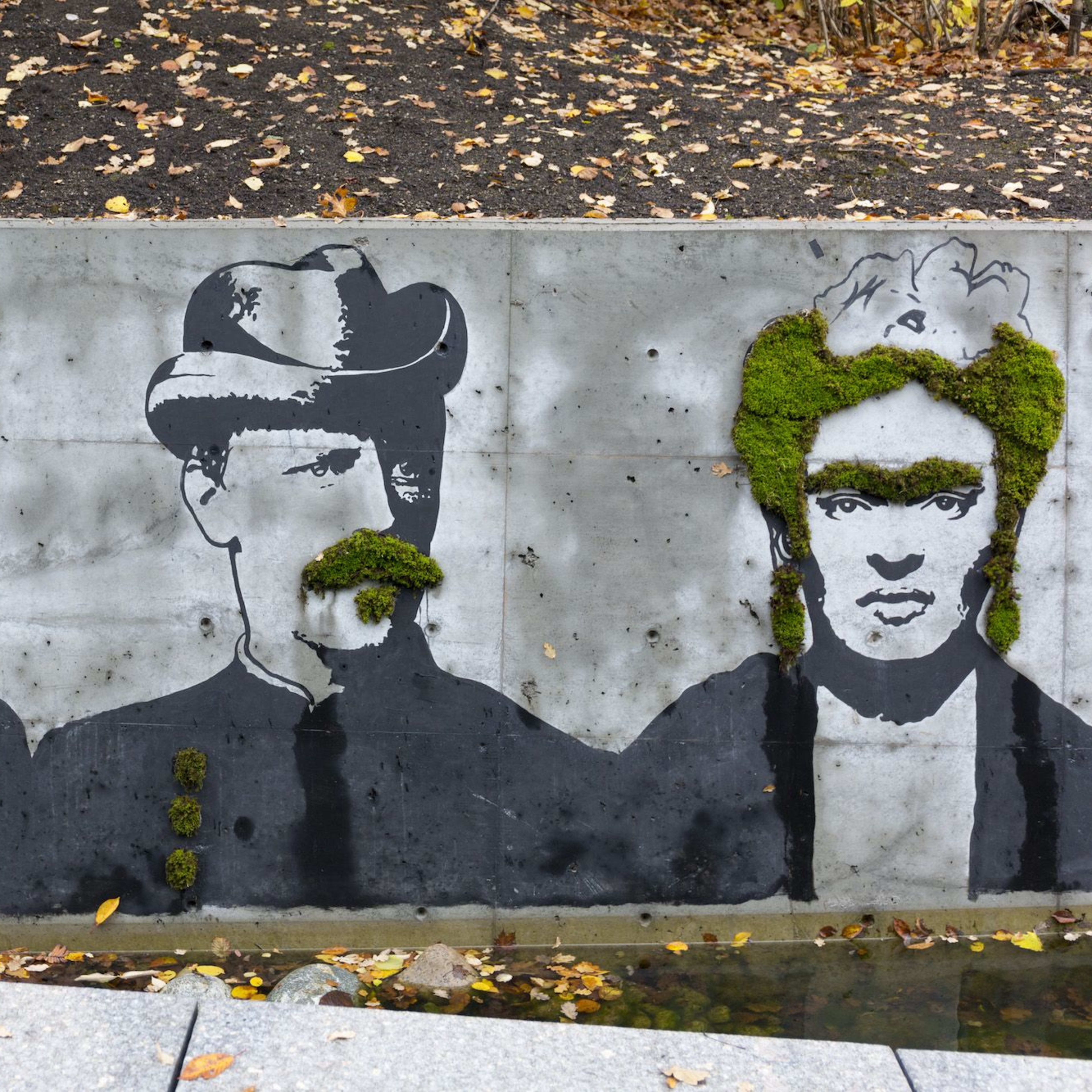 Straßenkunst an einer Betonwand mit drei schwarz-weißen Porträts, bei denen Moos als Haar und Bart verwendet wird.