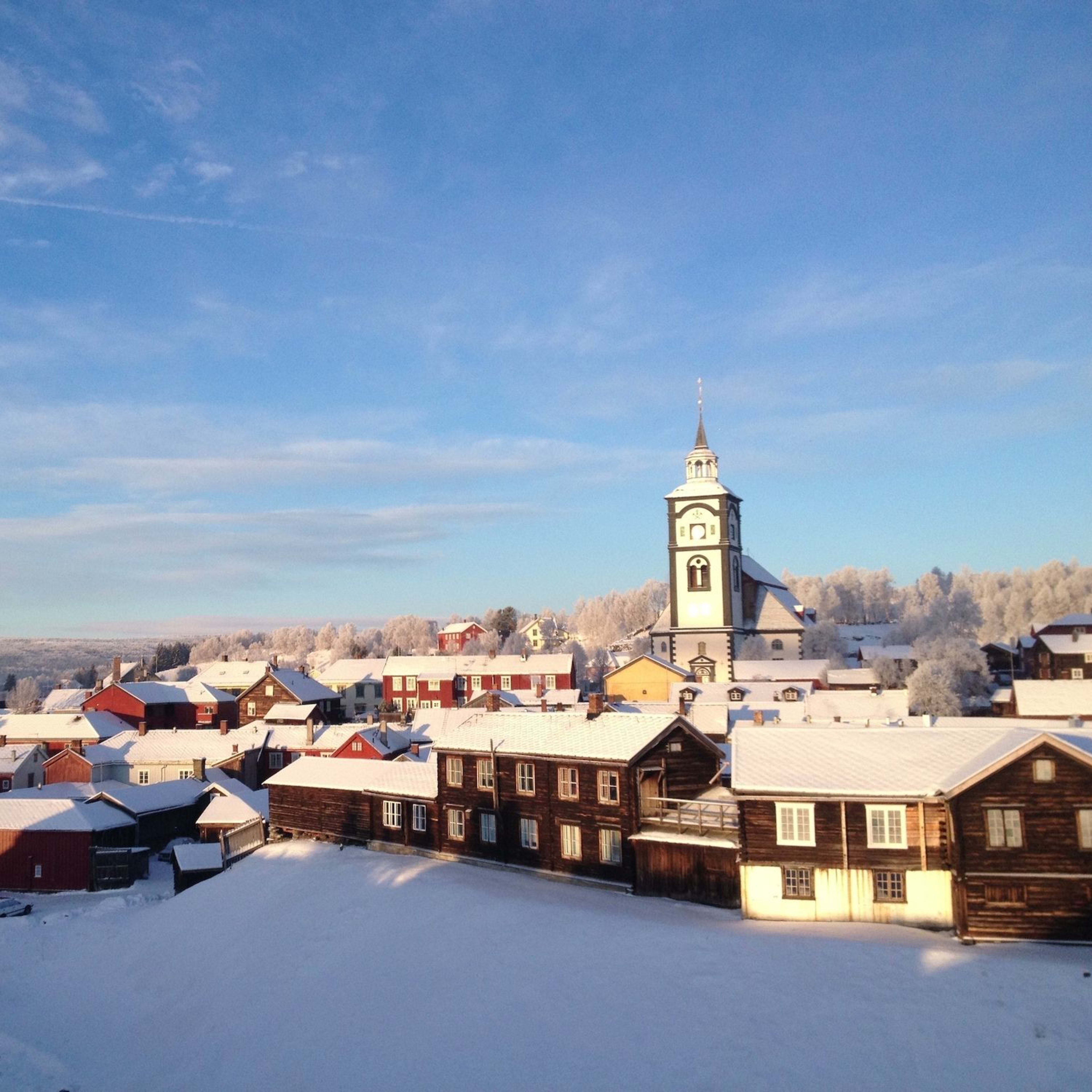 Snødekt landsby med fargerike trehus og høy kirketårn, omgitt av vinterlandskap under blå himmel.