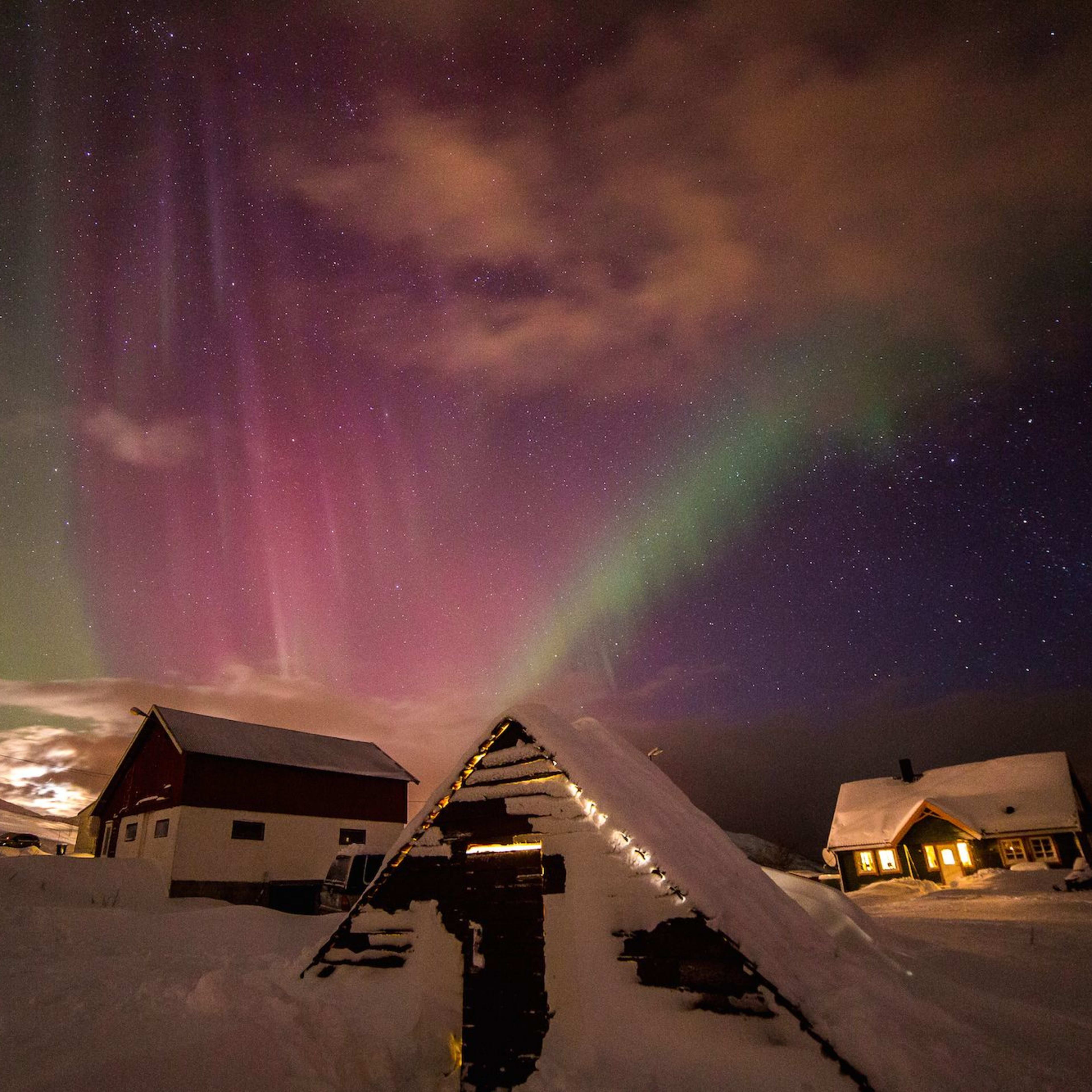Nordlys i grønne og rosa nyanser over snødekte hus, koselige hytter med varmt lys som skaper et magisk vintersceneri.