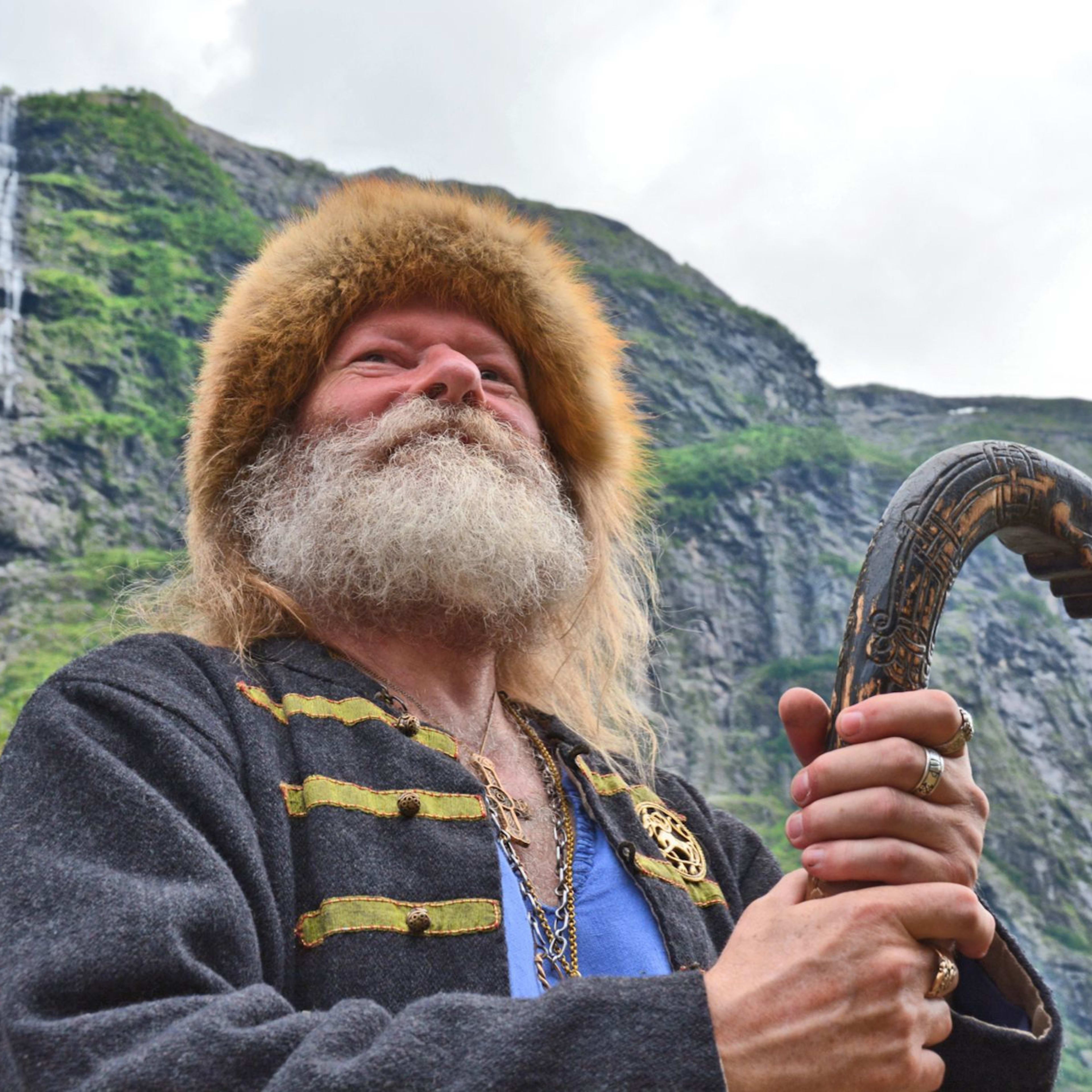 Bärtiger Mann mit Pelzmütze und traditioneller Kleidung hält einen geschnitzten Drachenstab, mit üppigen Bergen und einem rauschenden Wasserfall im Hintergrund.