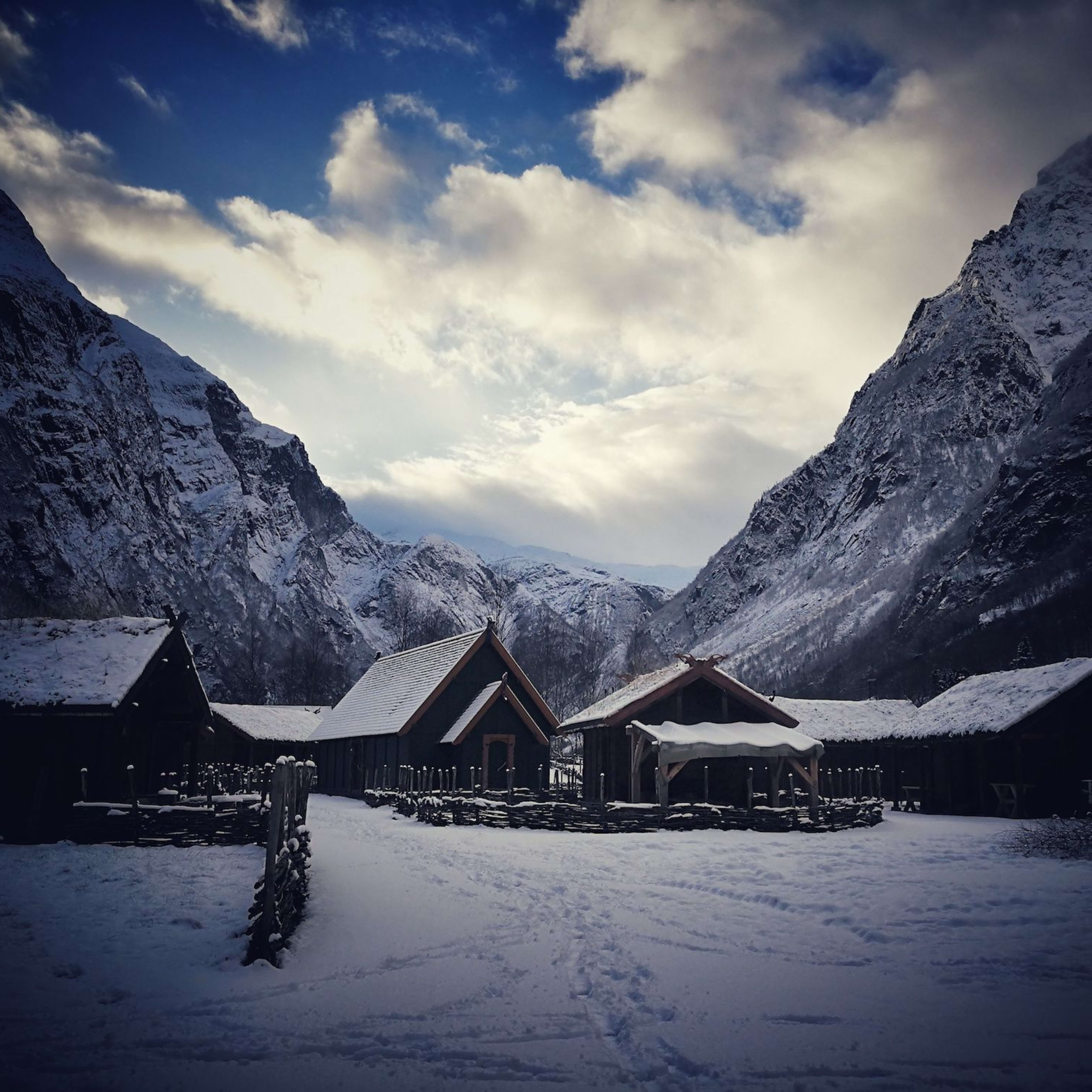 A snow-covered Viking village surrounded by towering mountains, with wooden houses blending into the winter landscape.