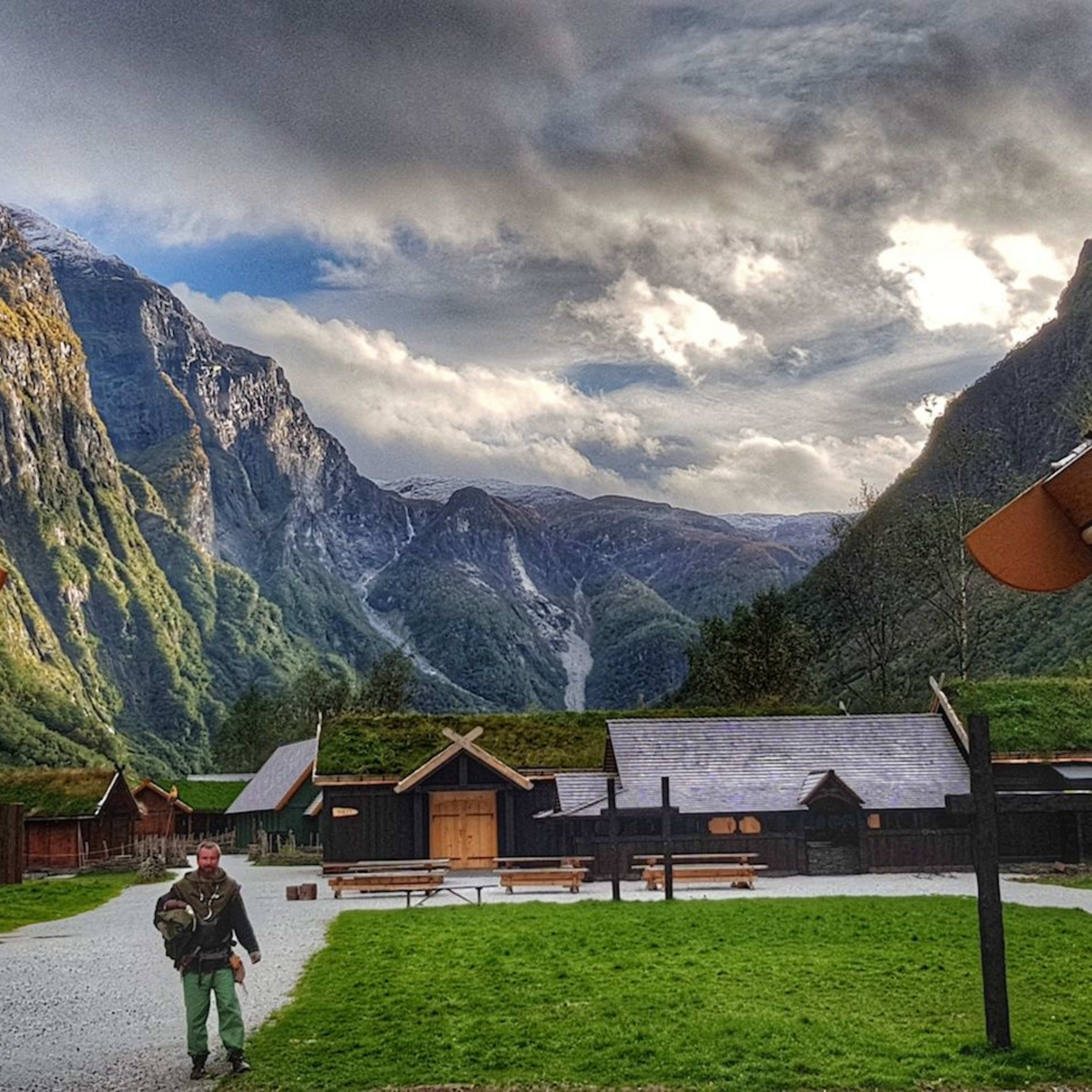 Ein Wikinger-Dorf mit grasbedeckten Dächern, umgeben von grünen Bergen unter dramatischem Himmel.