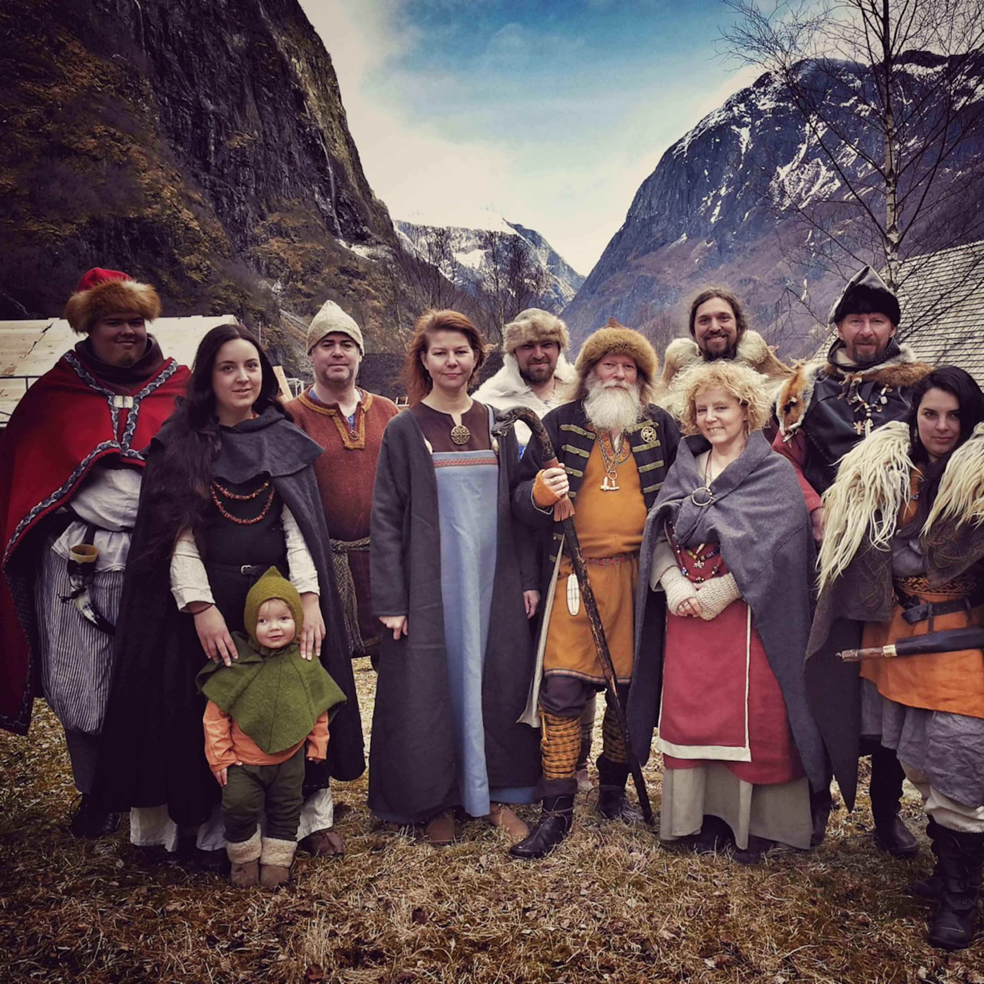Group dressed in Viking-style clothing, posing outdoors with snow-dusted mountains, cabins, and cloudy sky in the background.