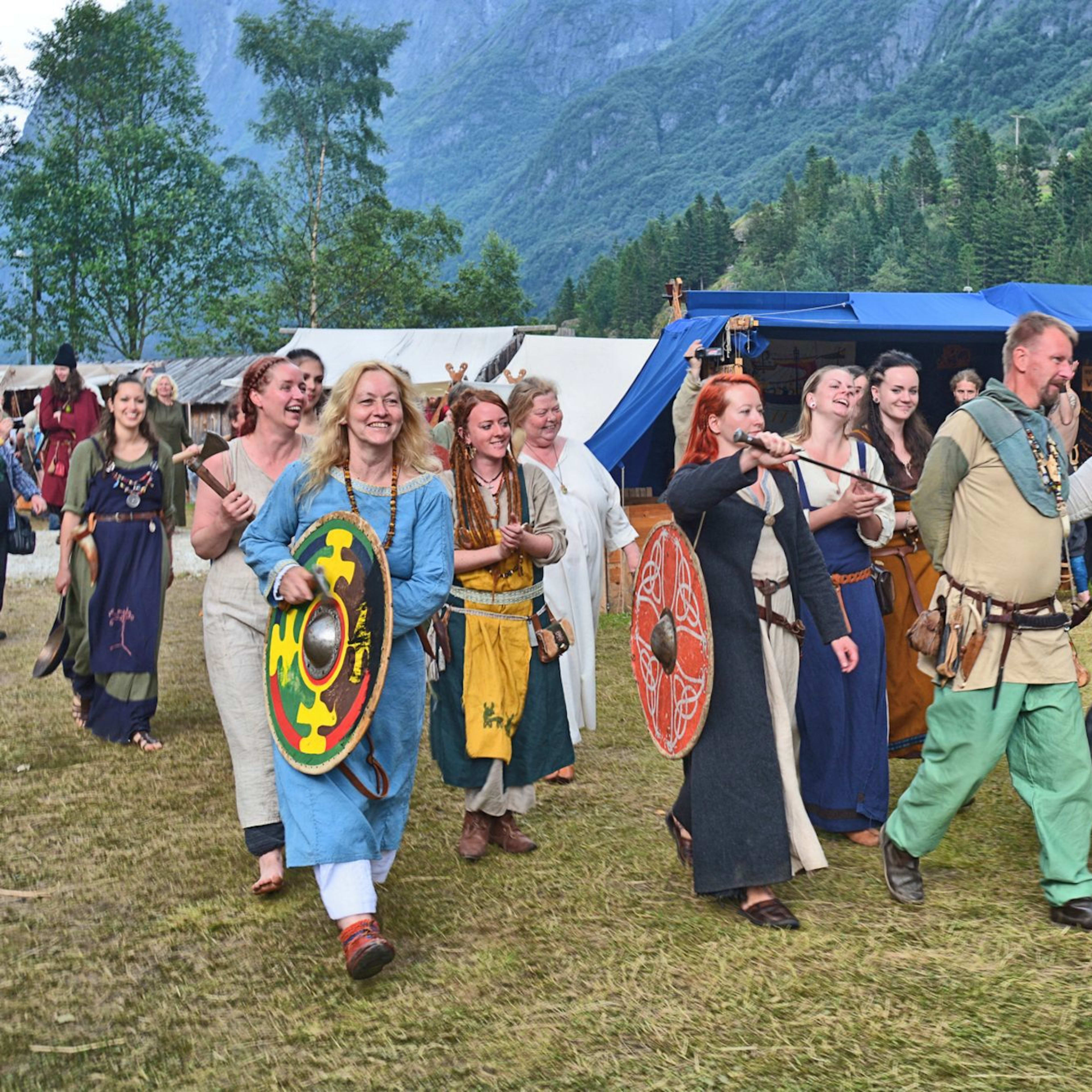 En livlig gruppe kledd som vikinger marsjerer gjennom en historisk rekonstruksjon, bærende skjold og staver.