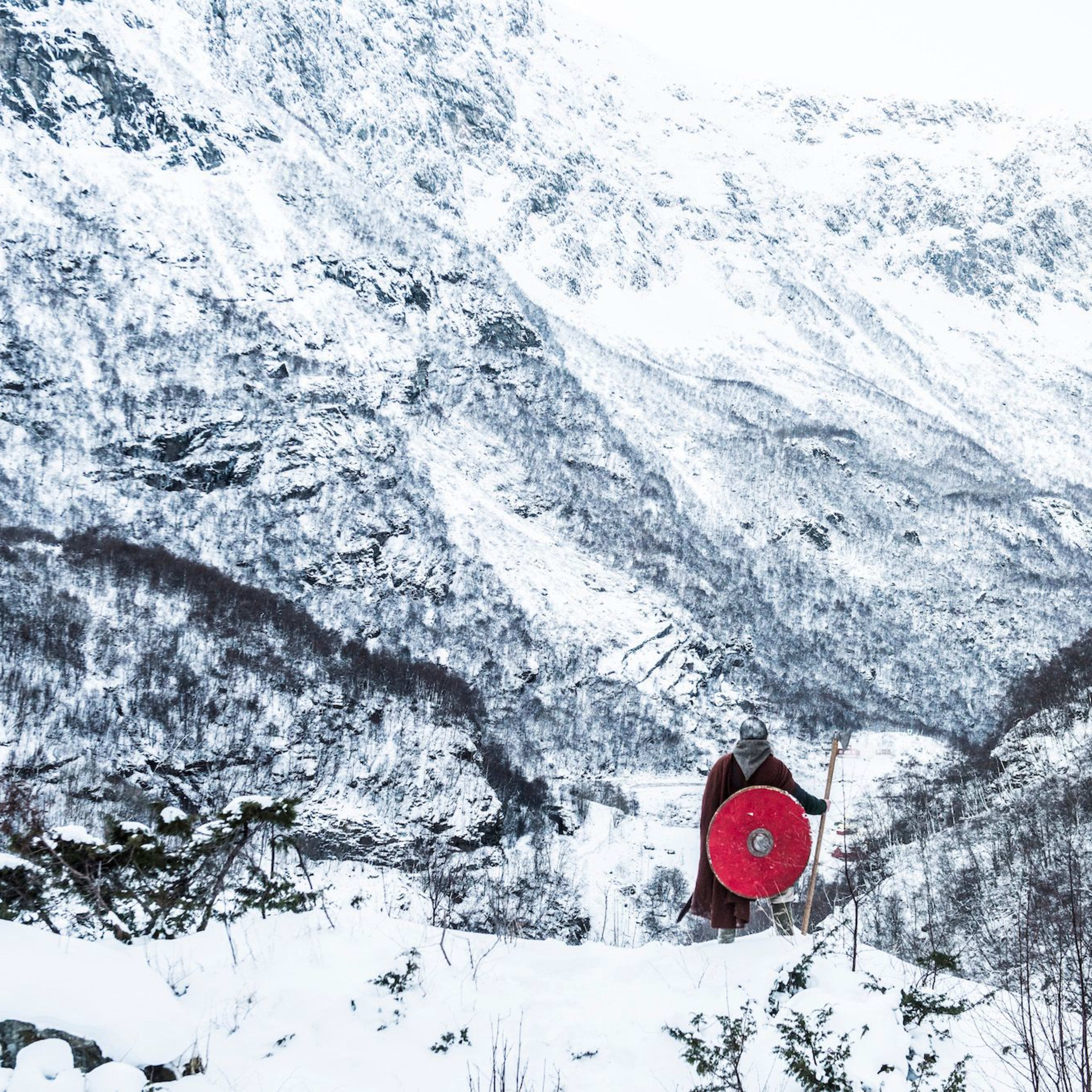 En viking i historisk antrekk på en snødekt fjellrygg med et rødt skjold og spyd.