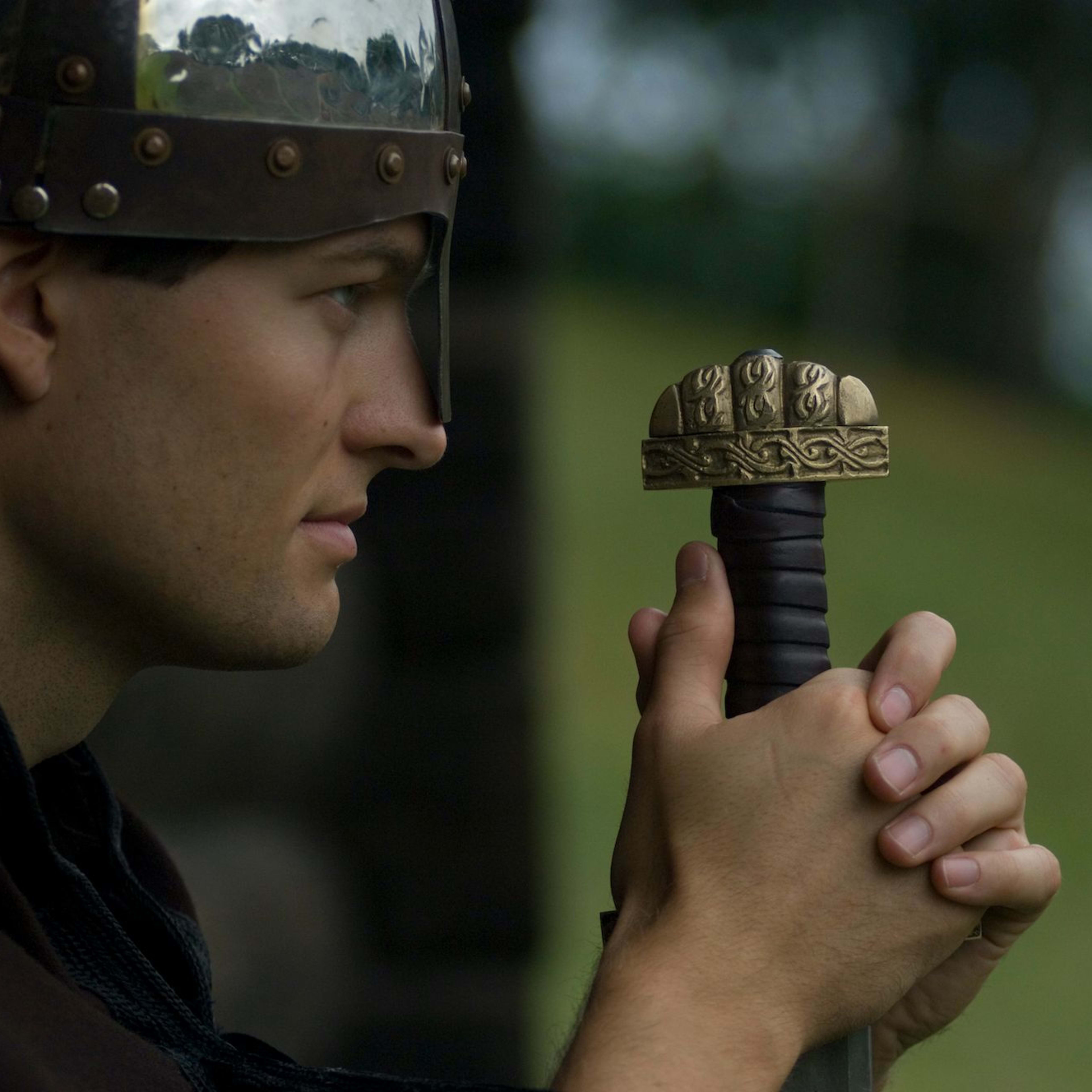 Seitenansicht eines Mannes mit einem Metallhelm, der ein Schwert mit komplizierten Gravuren hält.