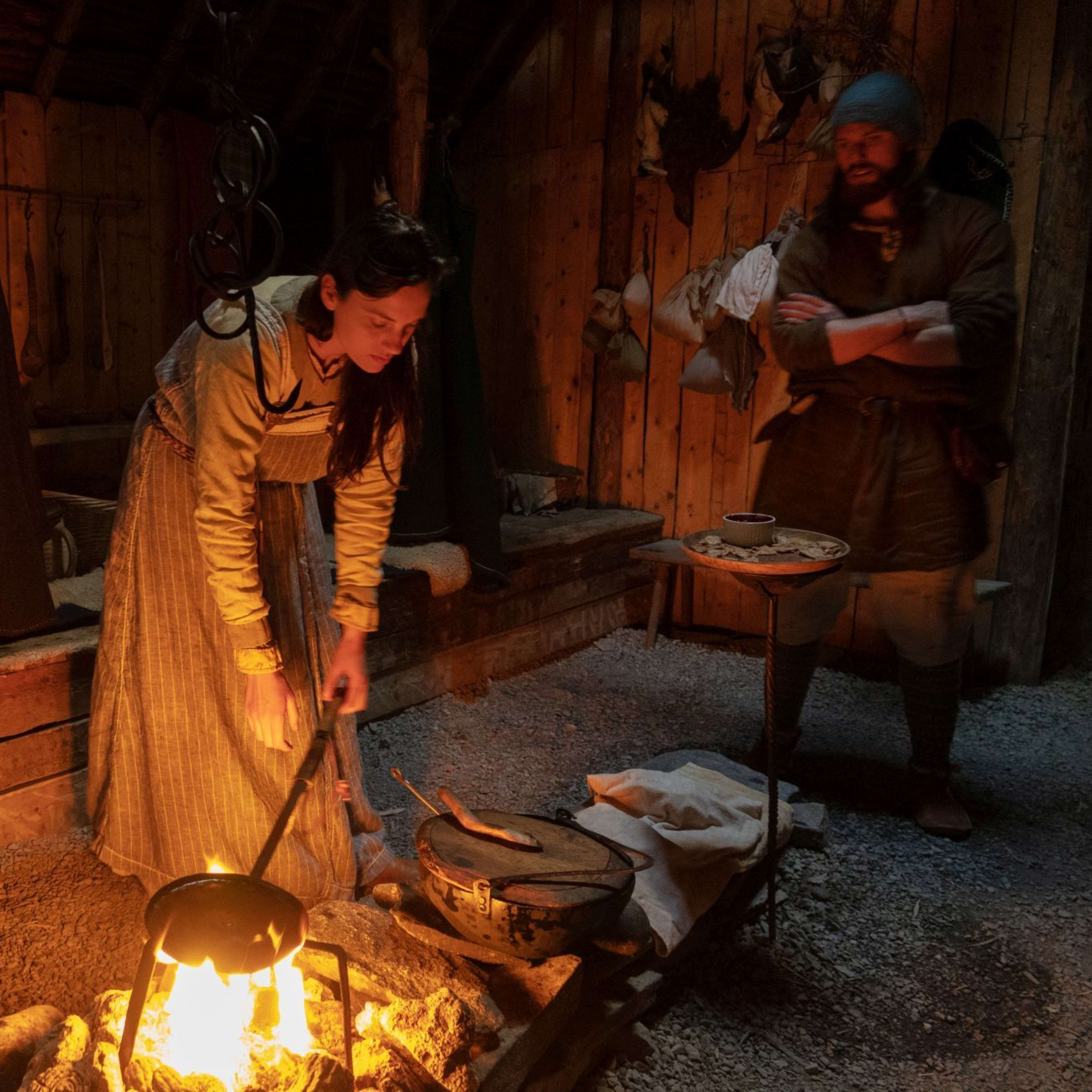Frau in traditioneller Kleidung, die einen Topf über offenem Feuer in einer rustikalen Hütte rührt, Mann schaut mit verschränkten Armen zu, gemütliches Interieur.