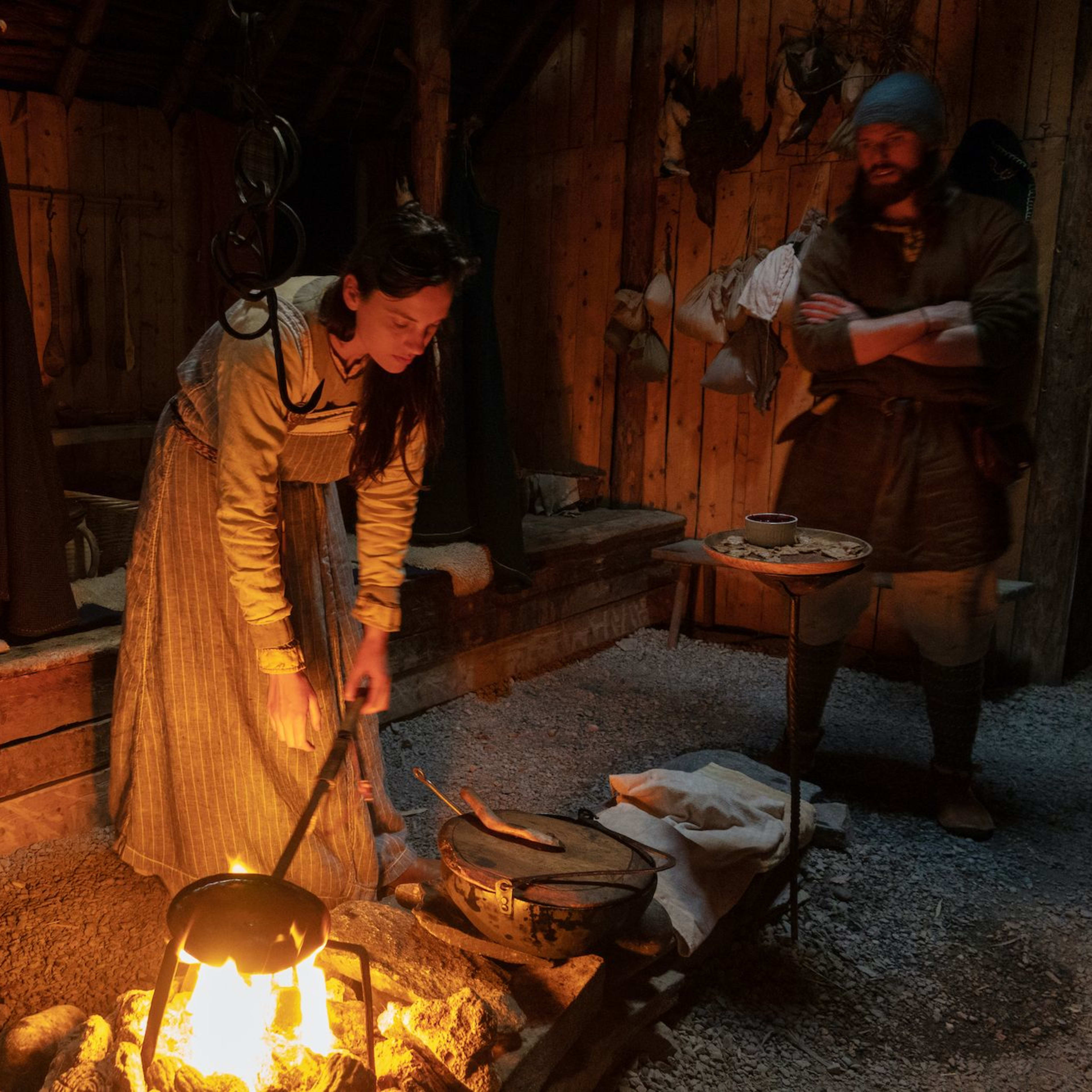 En kvinne i vikingtidsklær rører i en gryte over åpen ild i en svakt opplyst hytte.