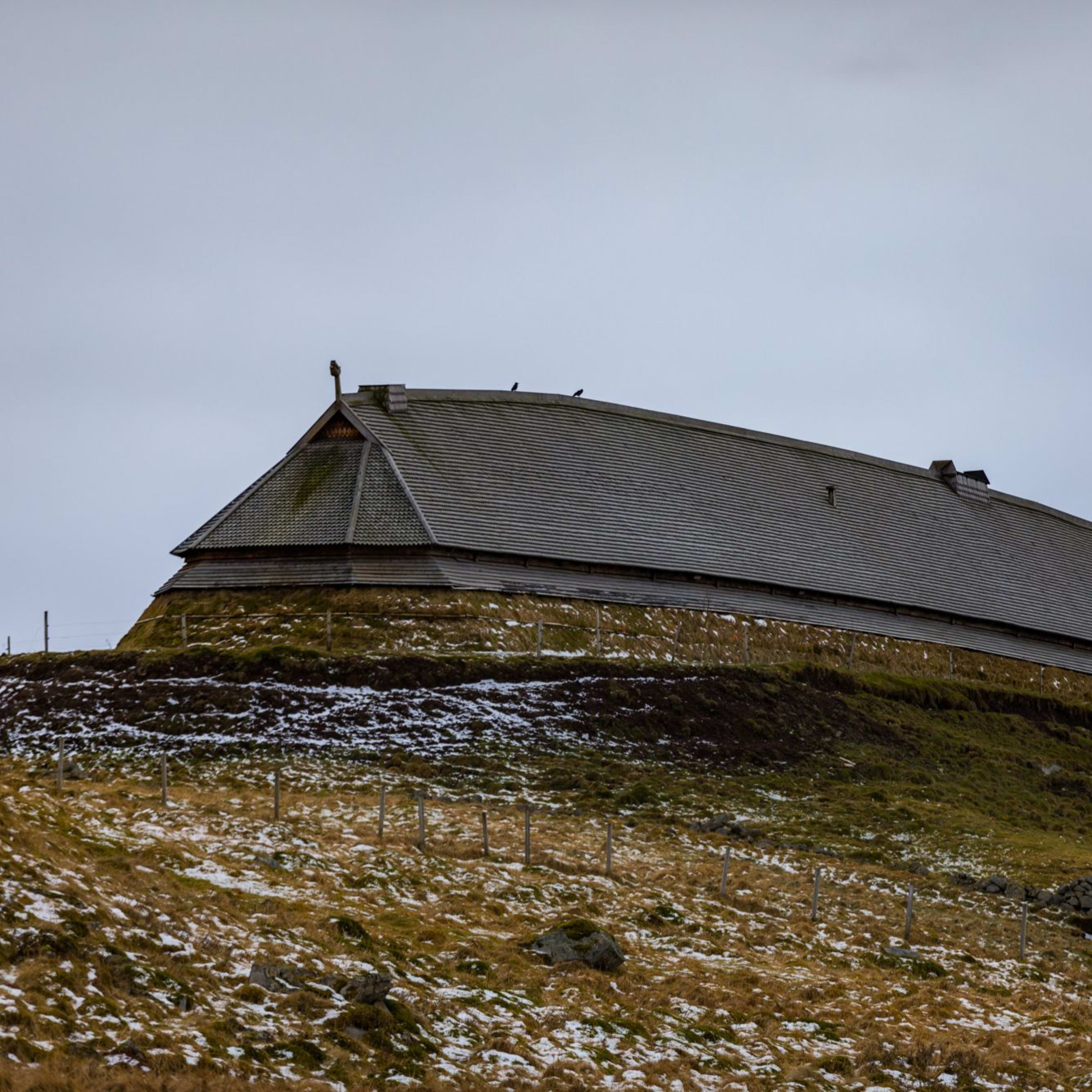 Wikinger-Langhaus auf einem grasbewachsenen Hügel mit Schneeflecken, Holzstruktur, bewölkter Himmel, der eine düstere, kalte Atmosphäre schafft.
