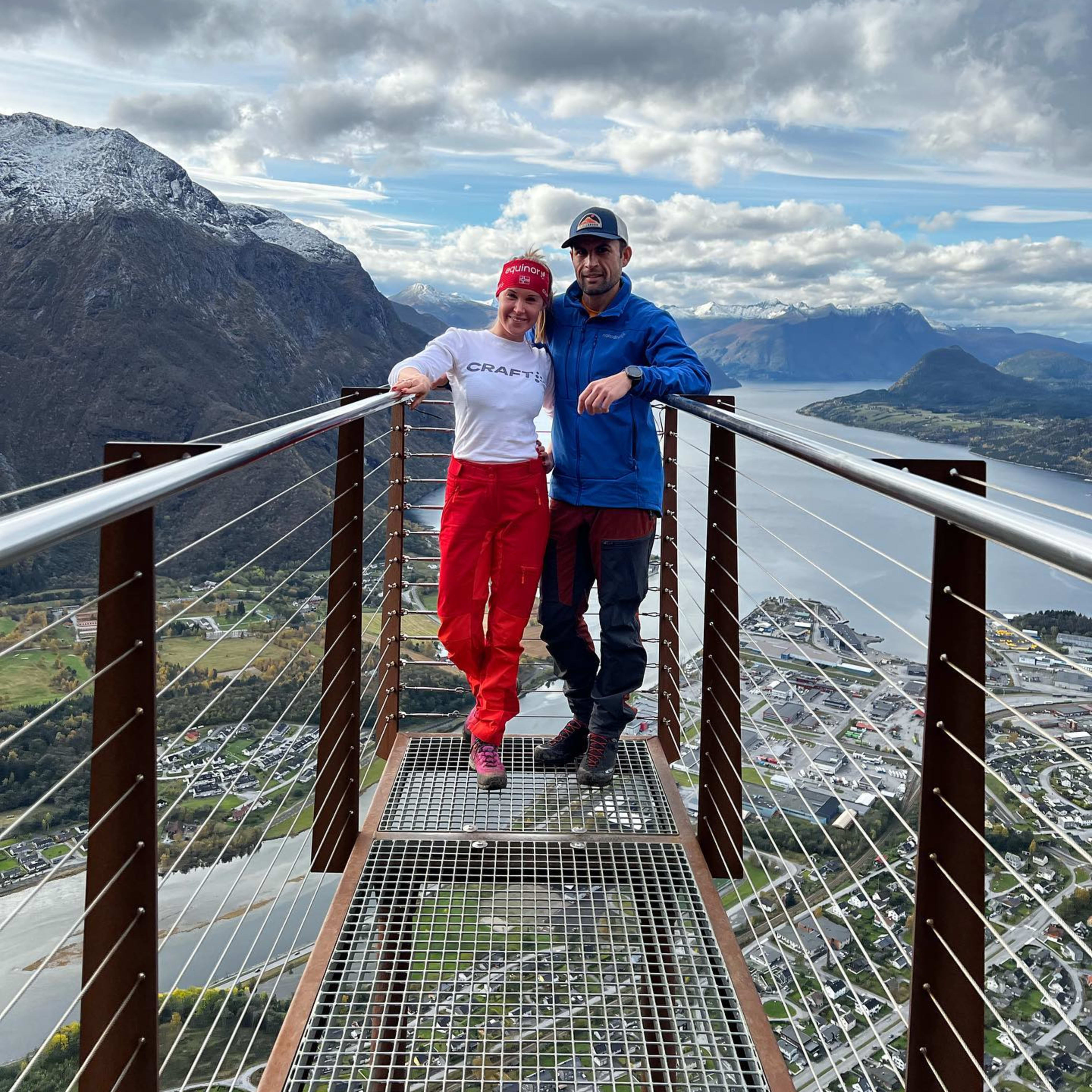 Par på utsiktsplattform med fjord og fjell i bakgrunnen under skyet himmel.