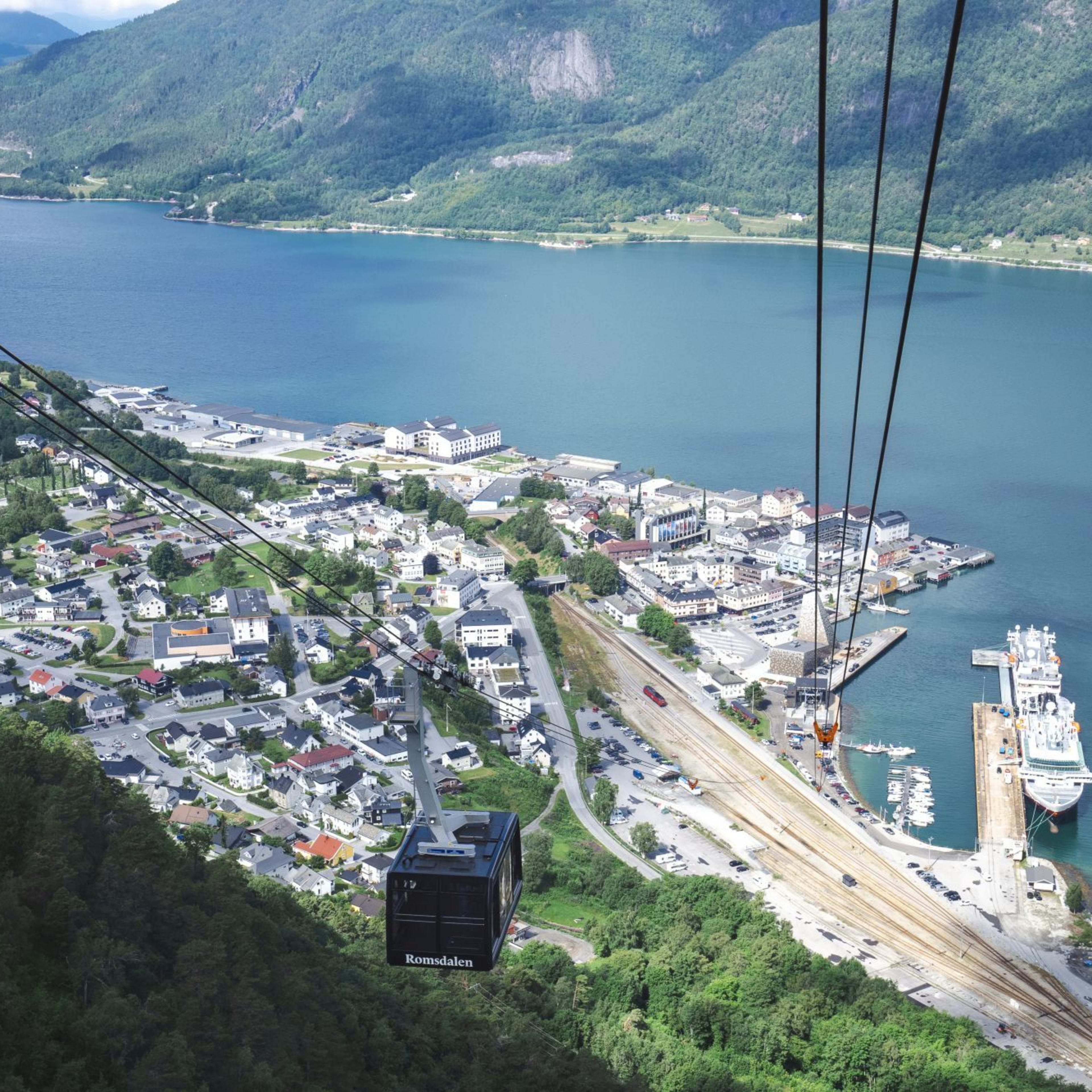 Luftfoto av kystby med taubane over skog og havn omgitt av fjell.