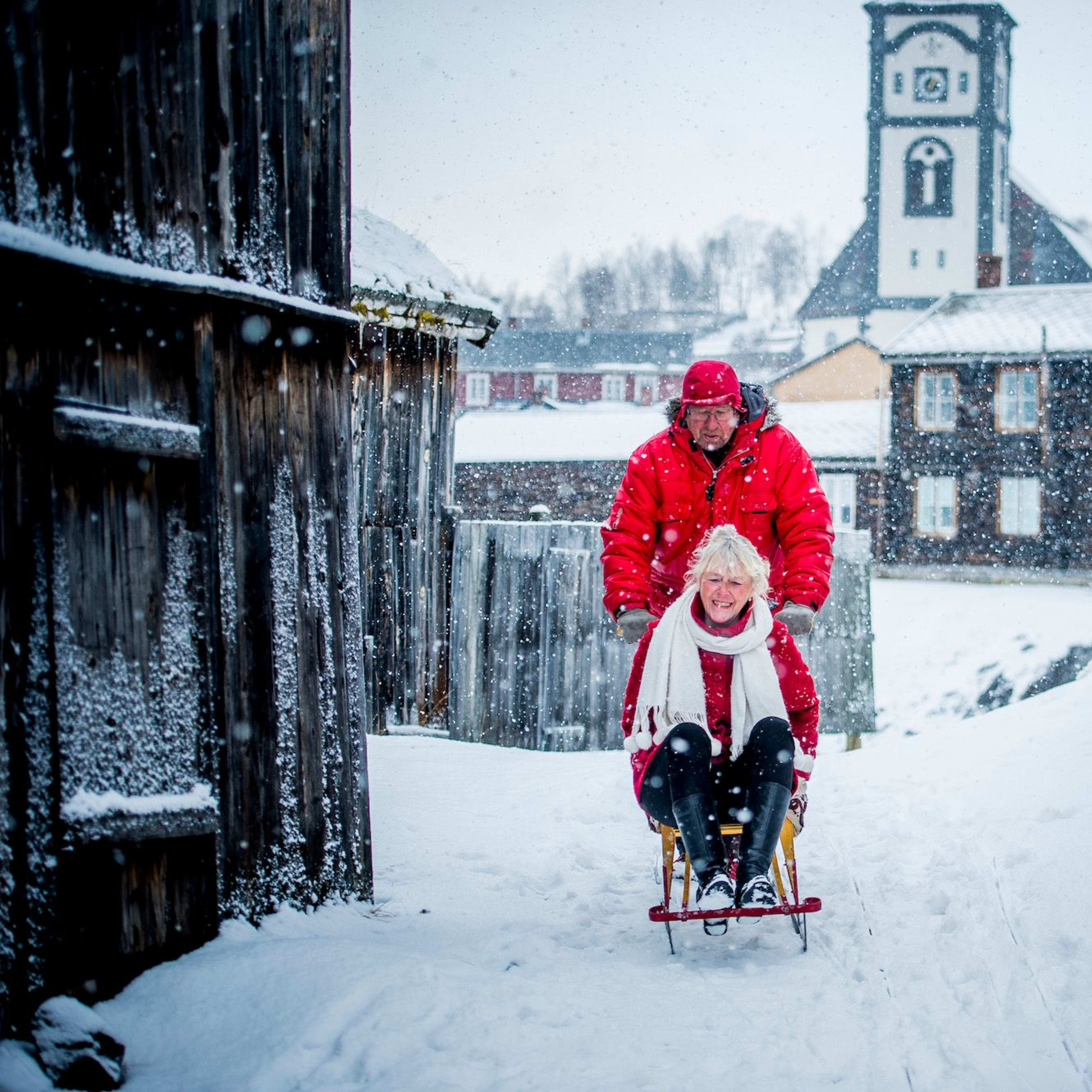 Eldre par i rødt nyter snøen, kvinne ler på kjelke mens mannen dytter.