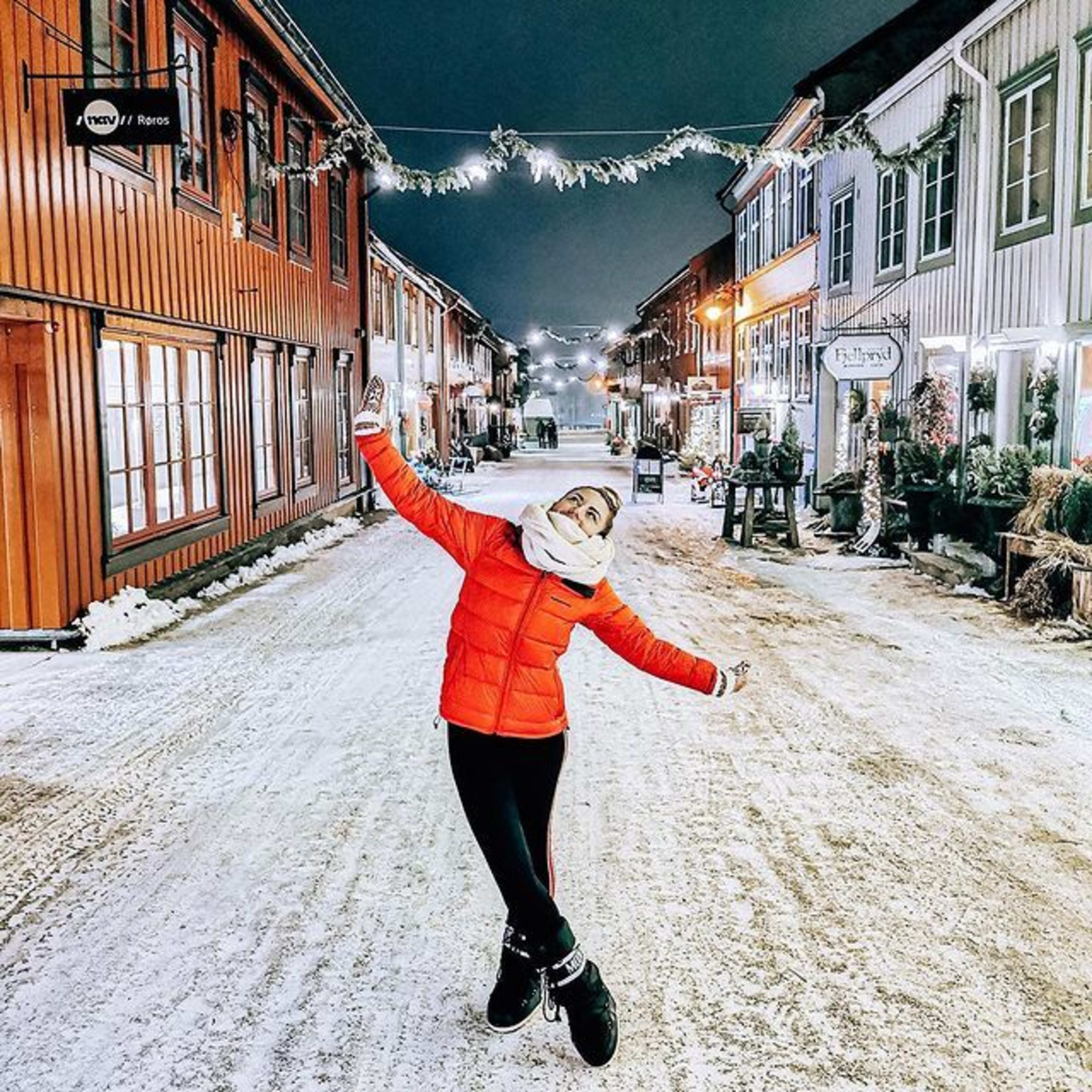 Kvinne i rødt smiler på snødekt gate med lys og fargerike hus.