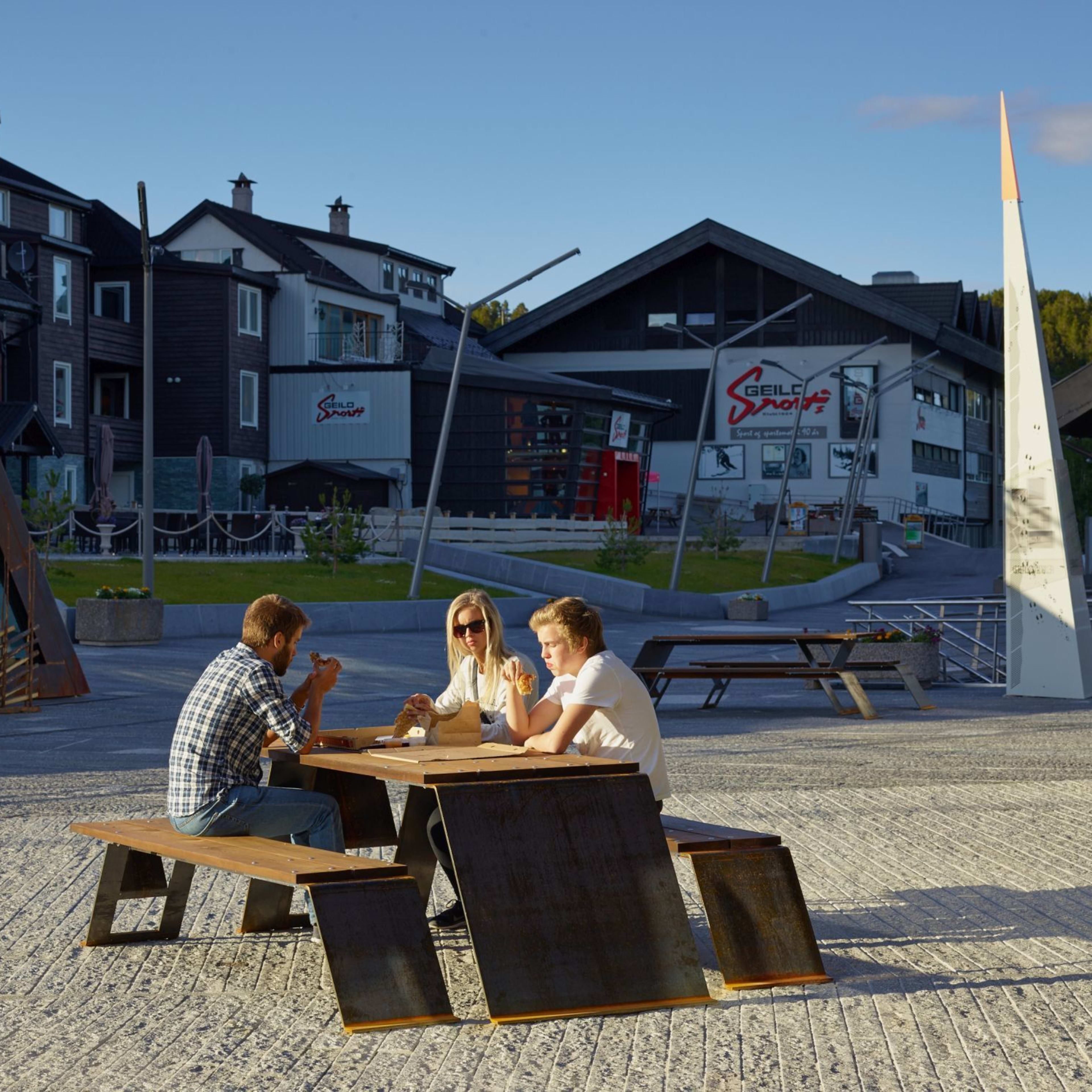 Drei essen Eis an Holztisch vor alpinen Geschäften bei Sonne.