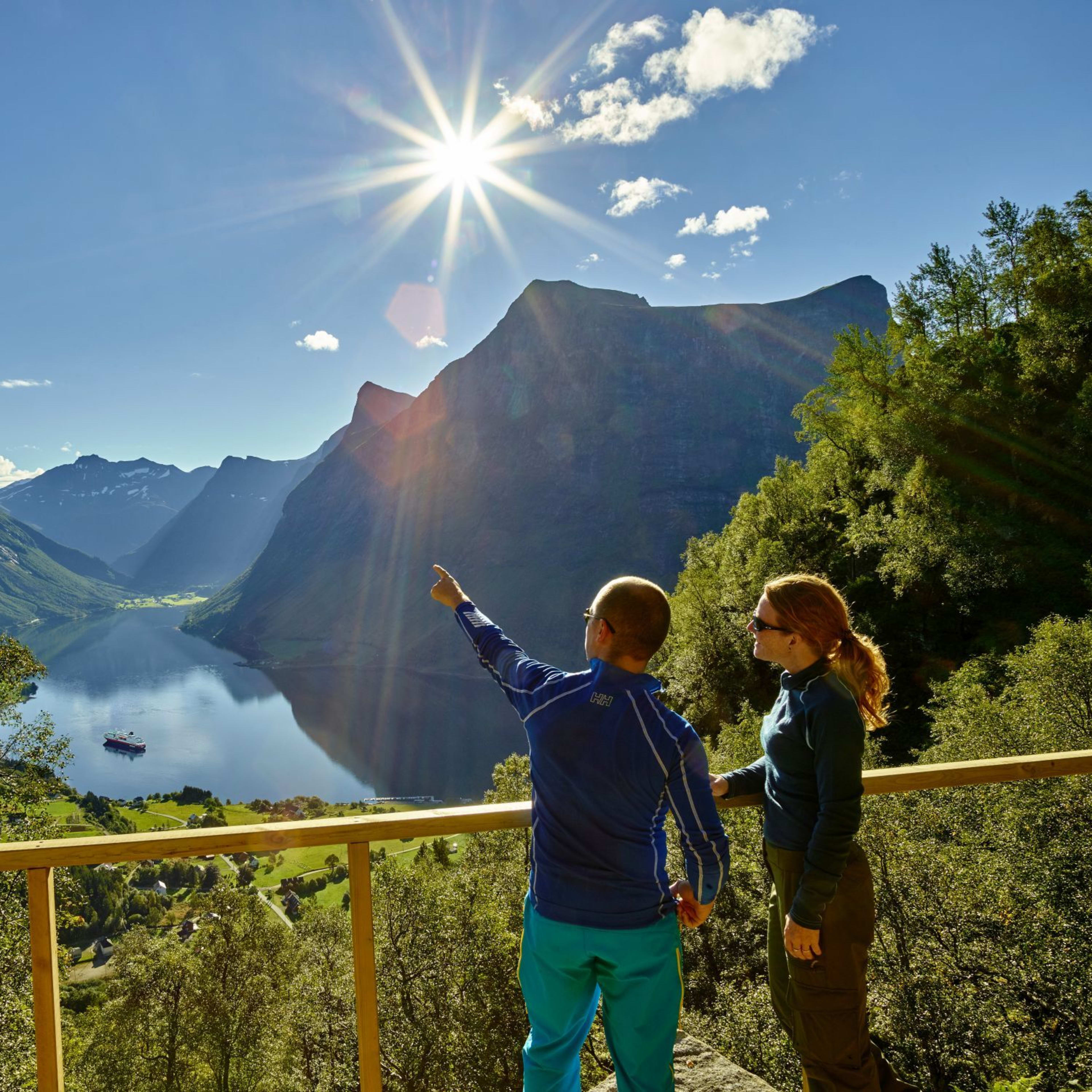 Par på utsiktsplattform som ser ut over norsk fjord, fjell, cruiseskip under og sol som kaster et varmt lys.