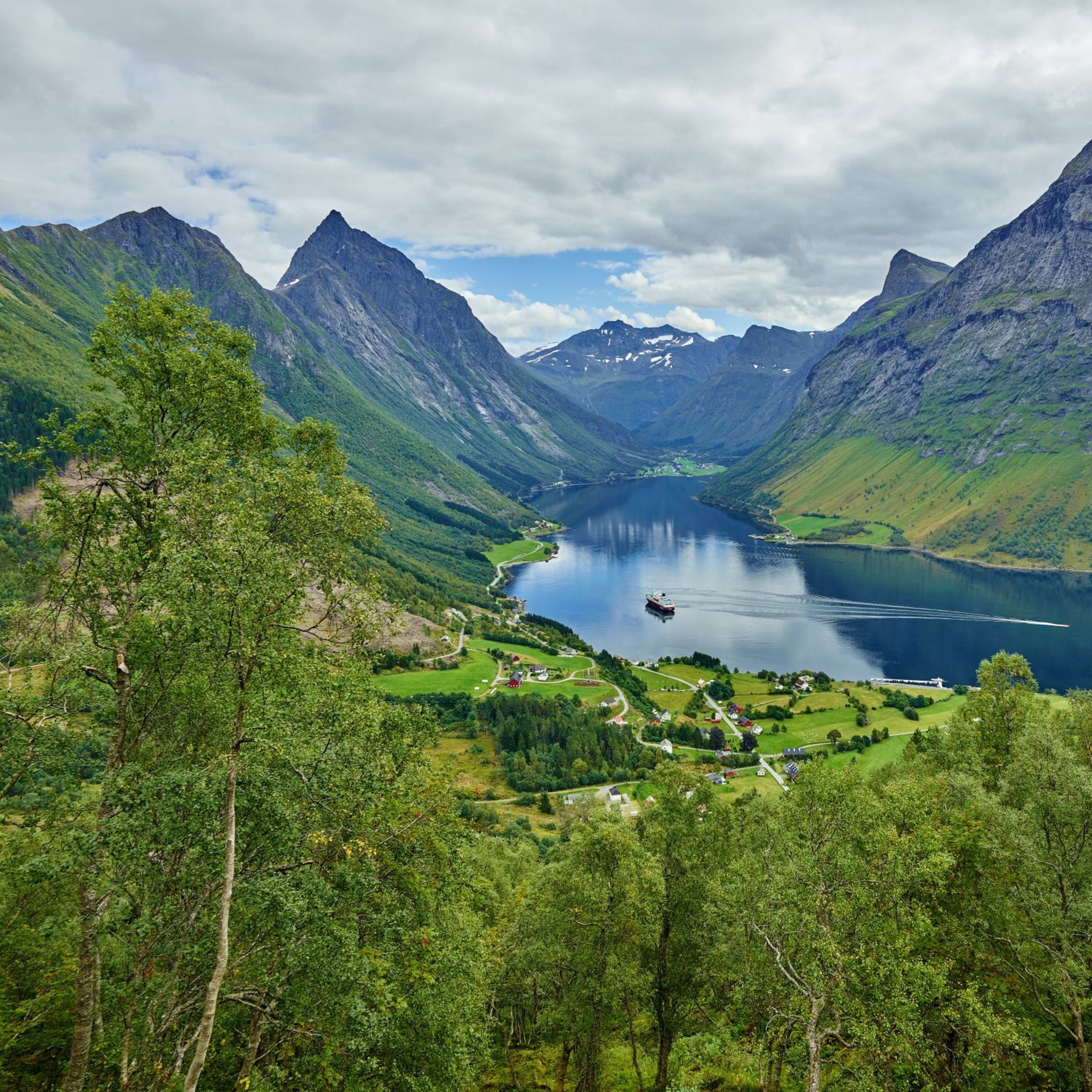 Scenic norsk fjord med liten cruiseskip, frodig grønt landskap, landsby langs vannkanten, snødekte topper og skyet himmel.