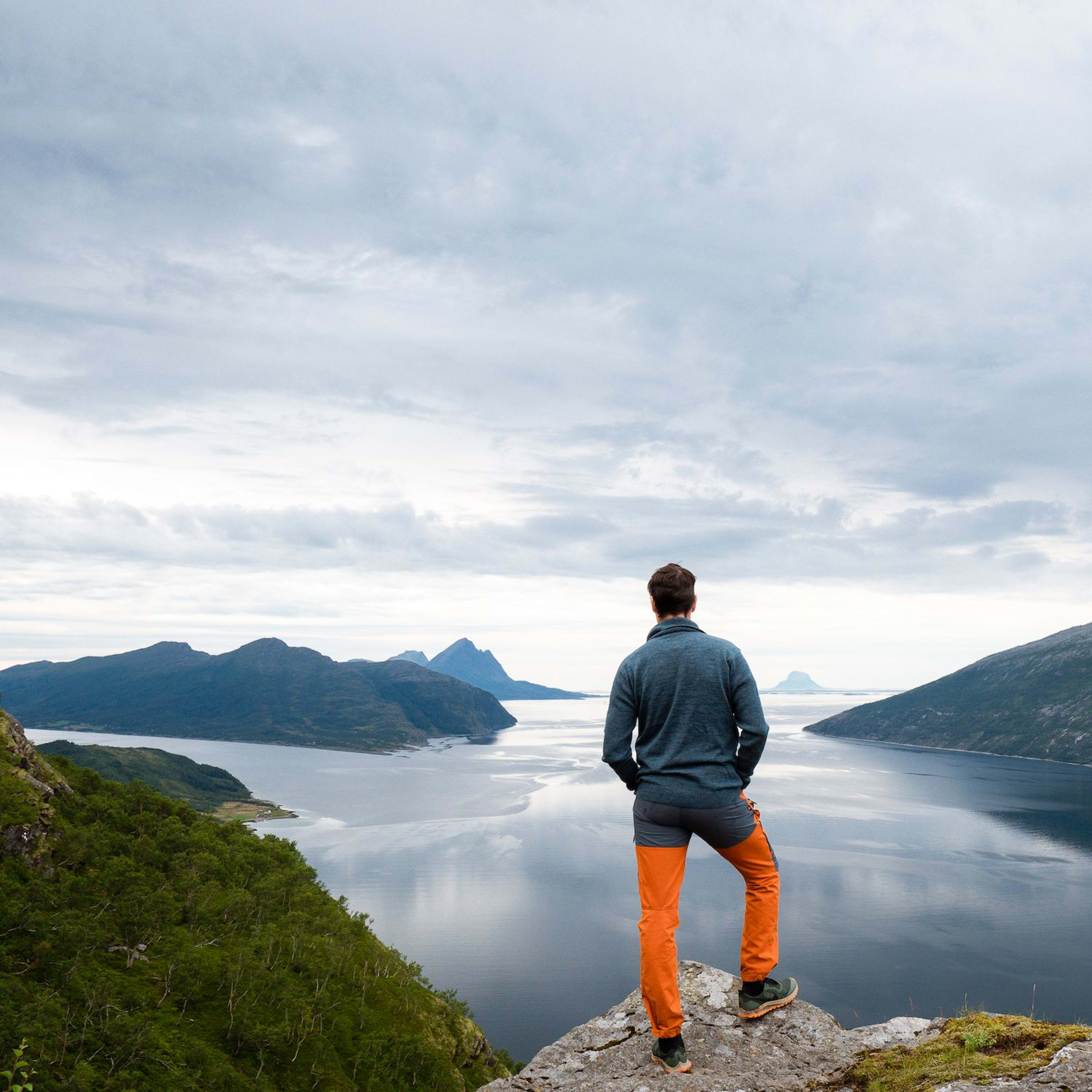 Person auf einem Felsen, der über den Fjord mit grünen Hügeln und fernen Bergen unter einem bewölkten Himmel blickt, was Abenteuer vermittelt.