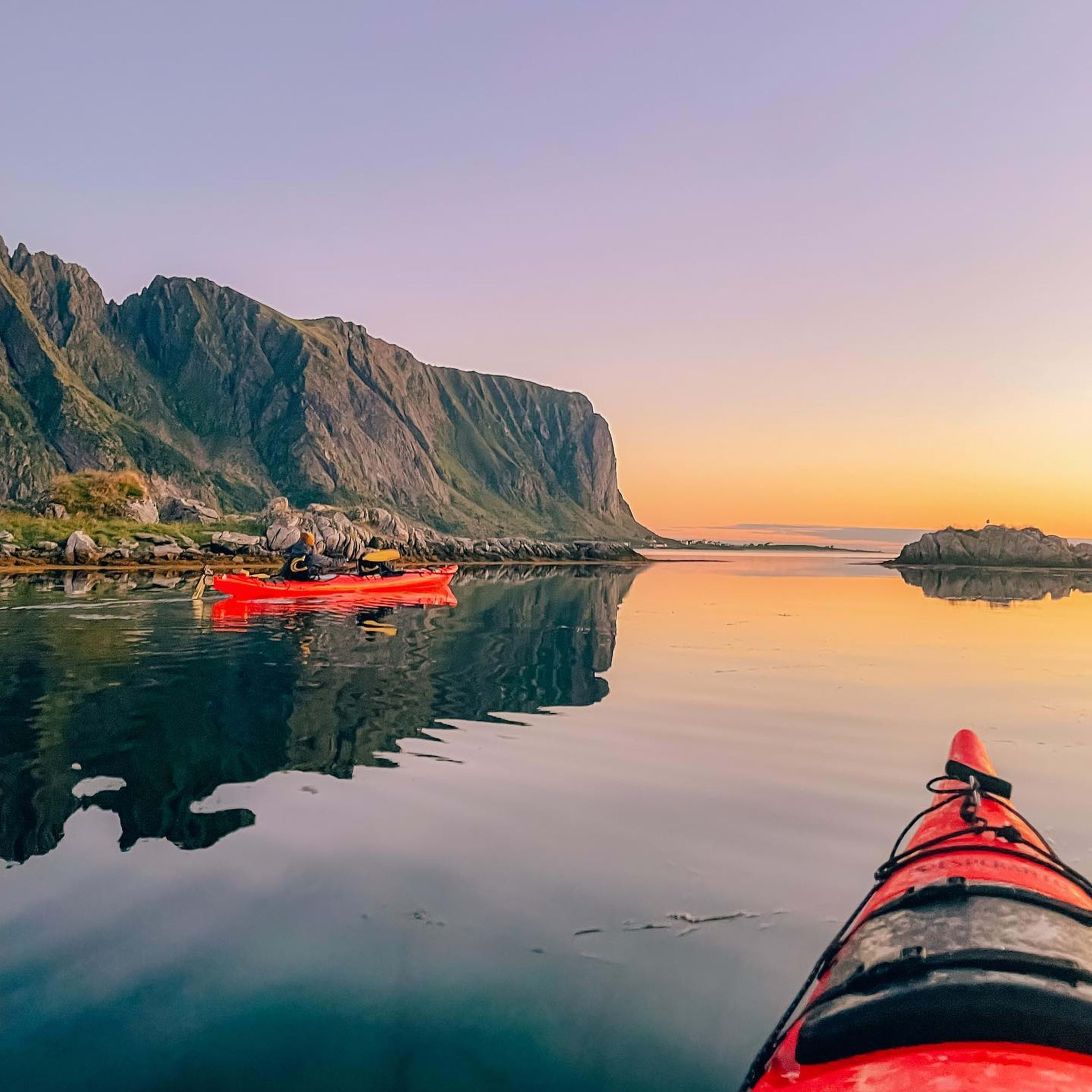 To røde kajakker padler i rolig fjord ved solnedgang med klipper og himmel speilet i vannet.