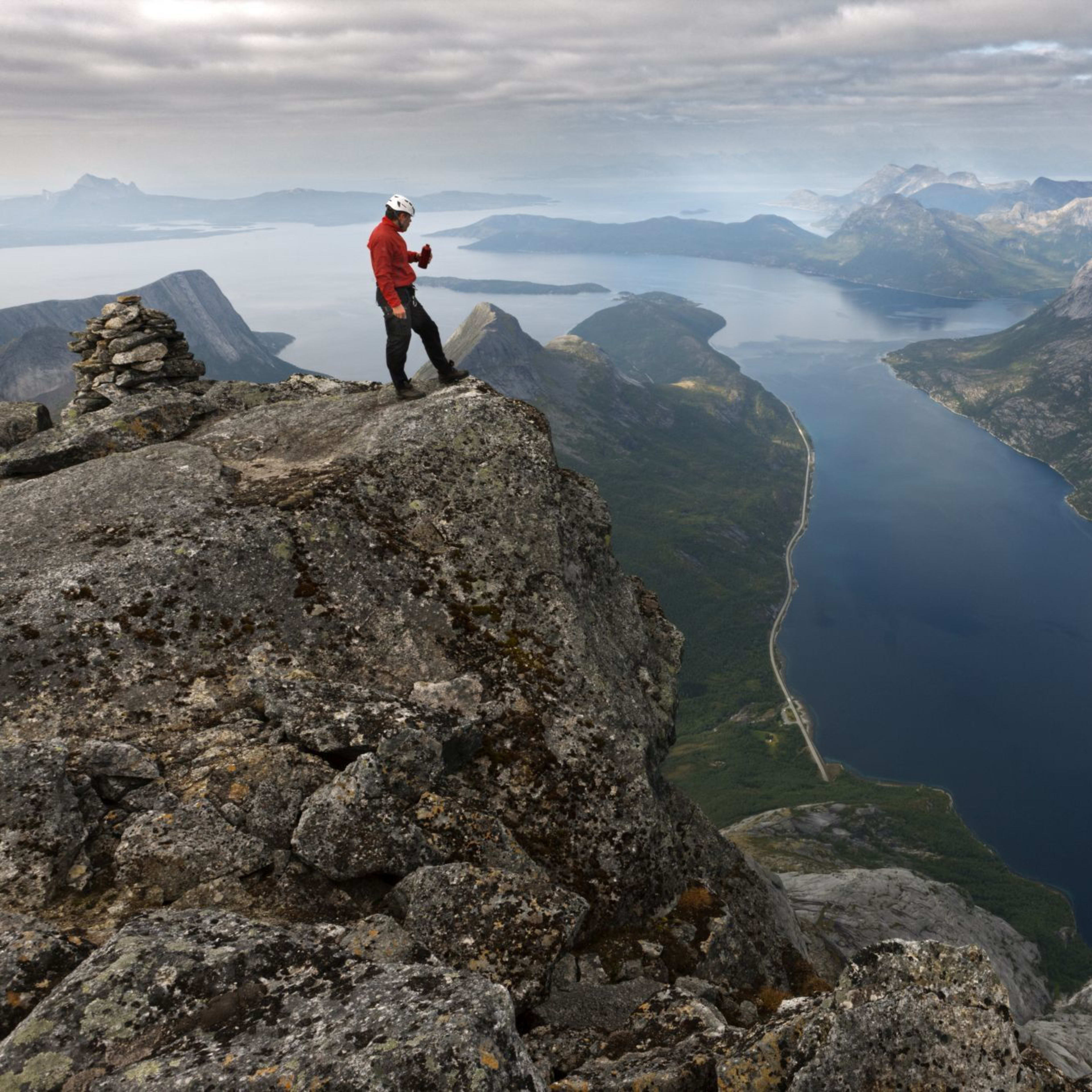 Hiker in red and black gear standing on cliff edge overlooking fjord with blue water, rugged mountains, and cloudy sky.