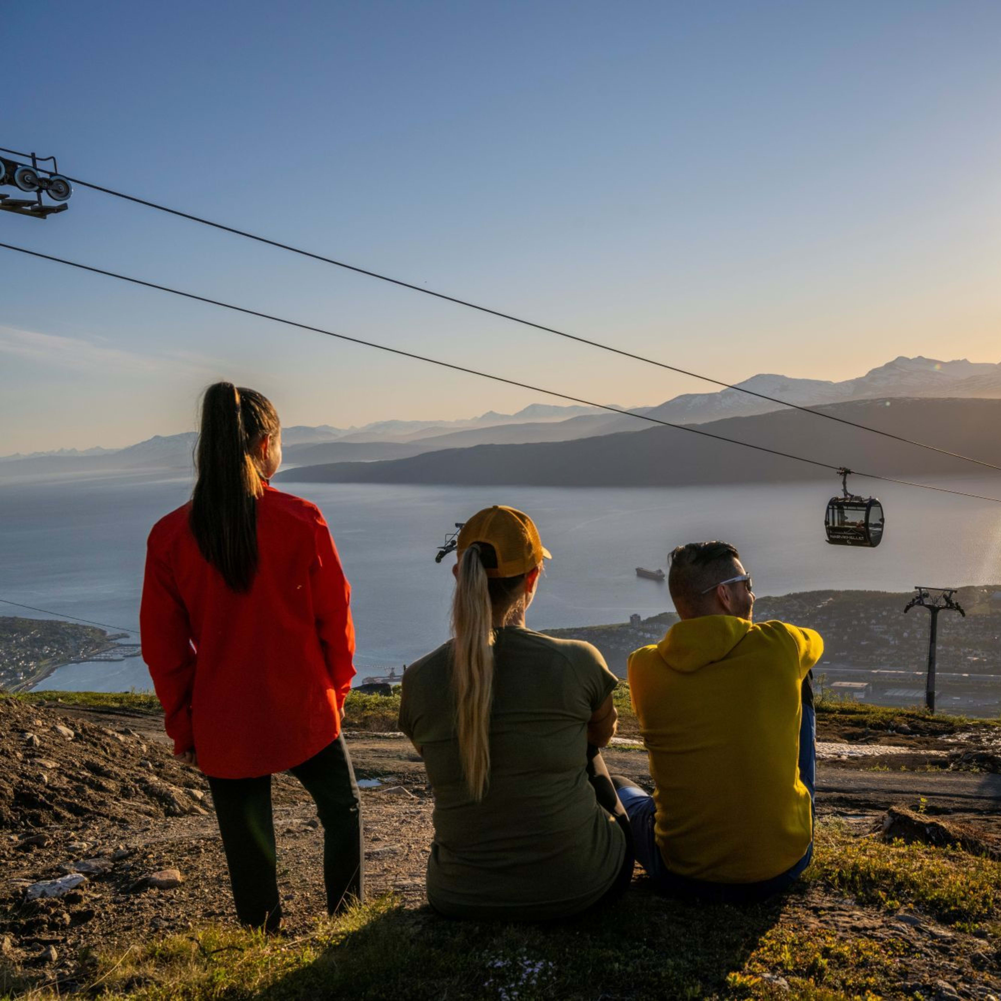 Three people sitting on hillside overlooking fjord with golden sunlight at sunset, two cable cars above and snow-capped mountains.