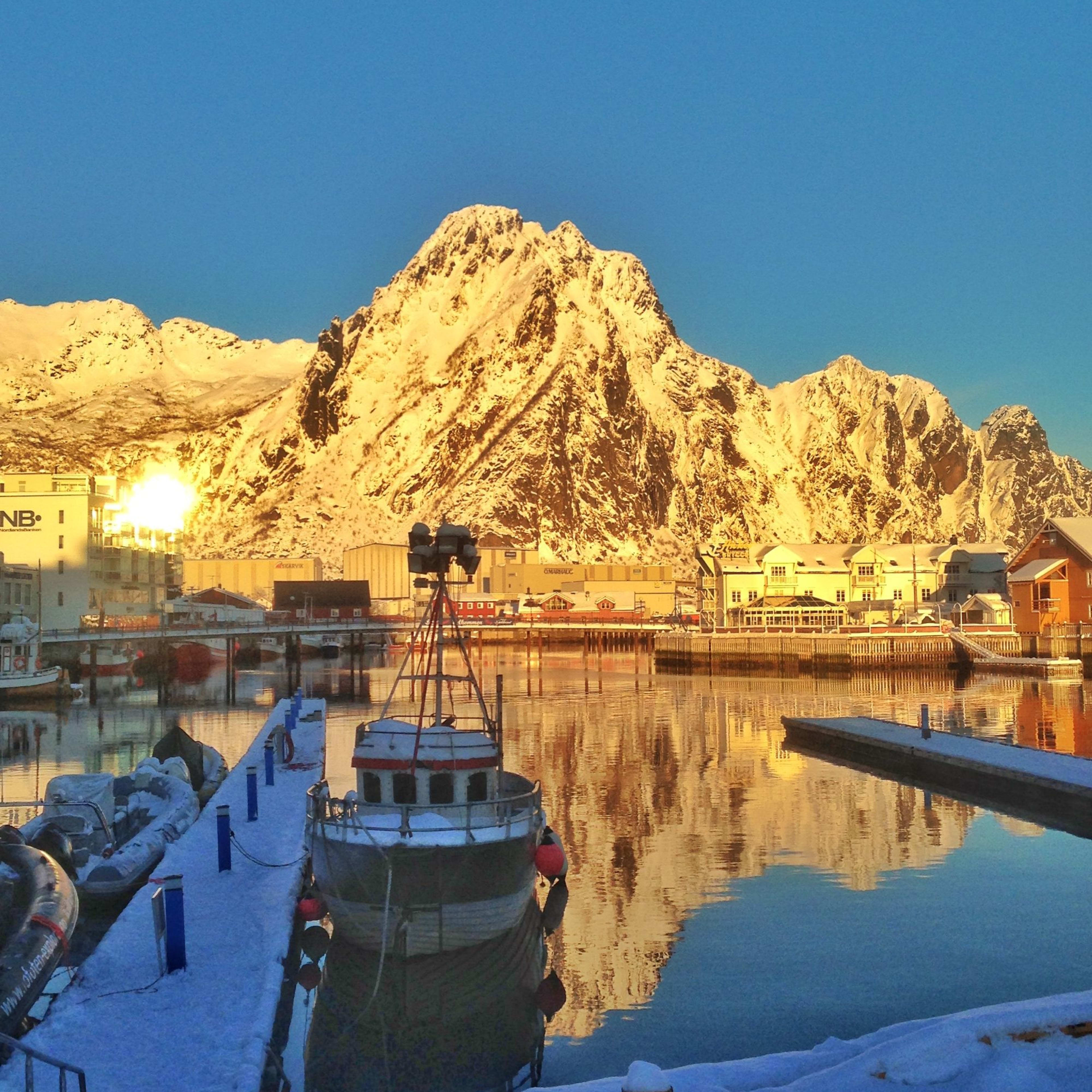 Båt i snødekt havn ved solnedgang, gyllent lys på fjell.