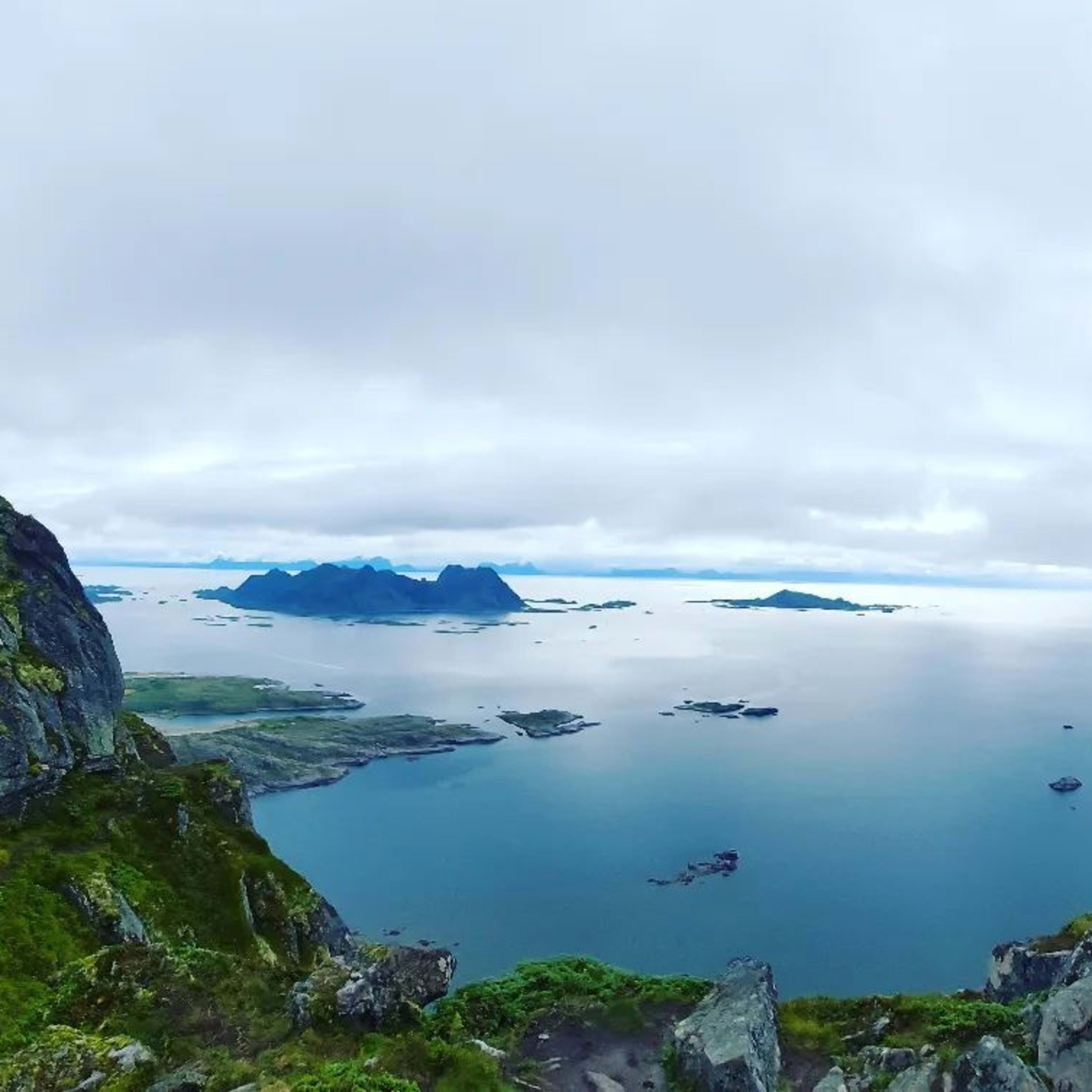 Kvinne på fjellkam ser ut over øyer og hav under grå himmel.