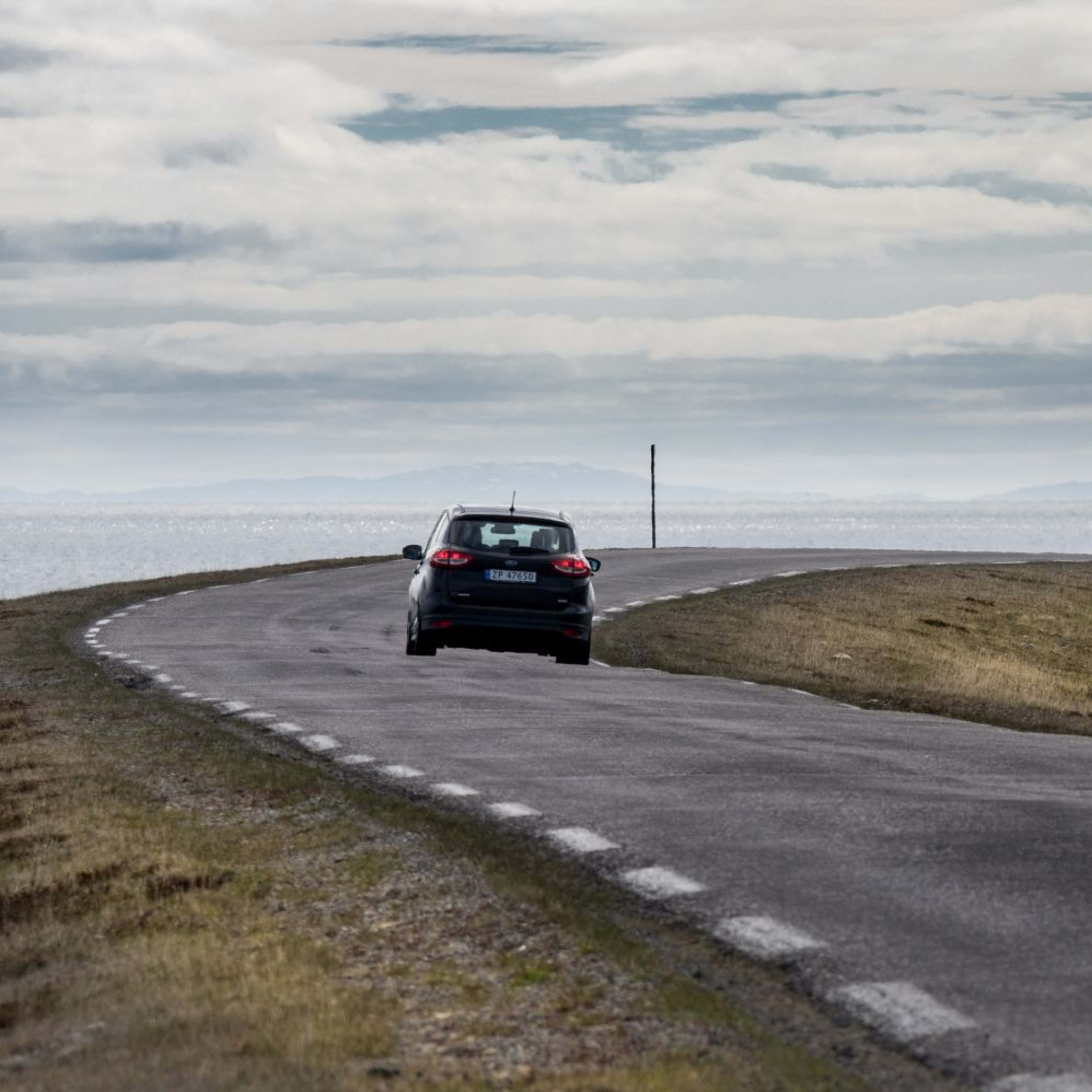 Ein einsames Auto fährt auf einer kurvigen Straße durch ödes Grasland und felsiges Terrain zum weiten Meer unter einem bewölkten Himmel.