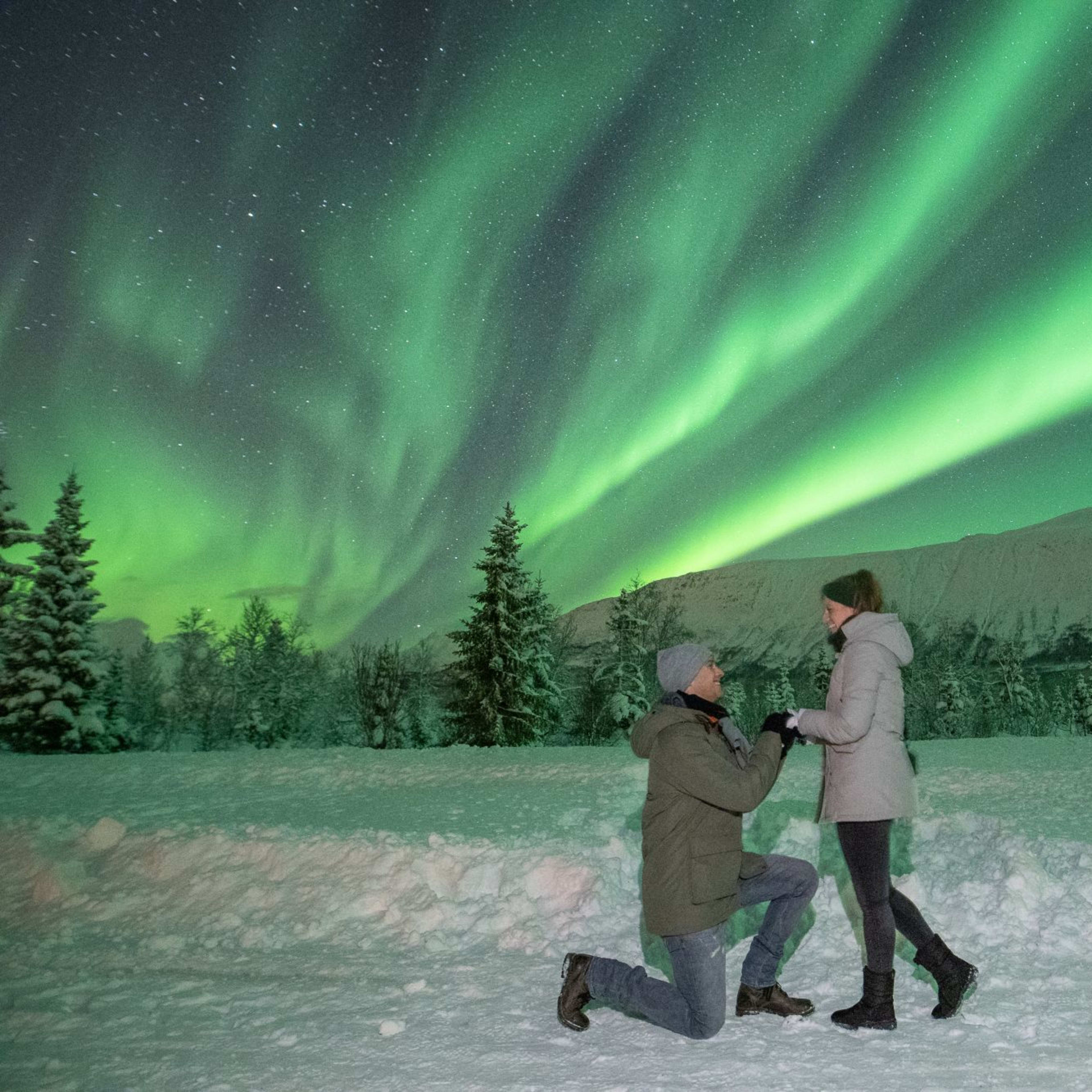 Romantic winter scene with couple under Northern Lights, one proposing on knee in snow-covered landscape.