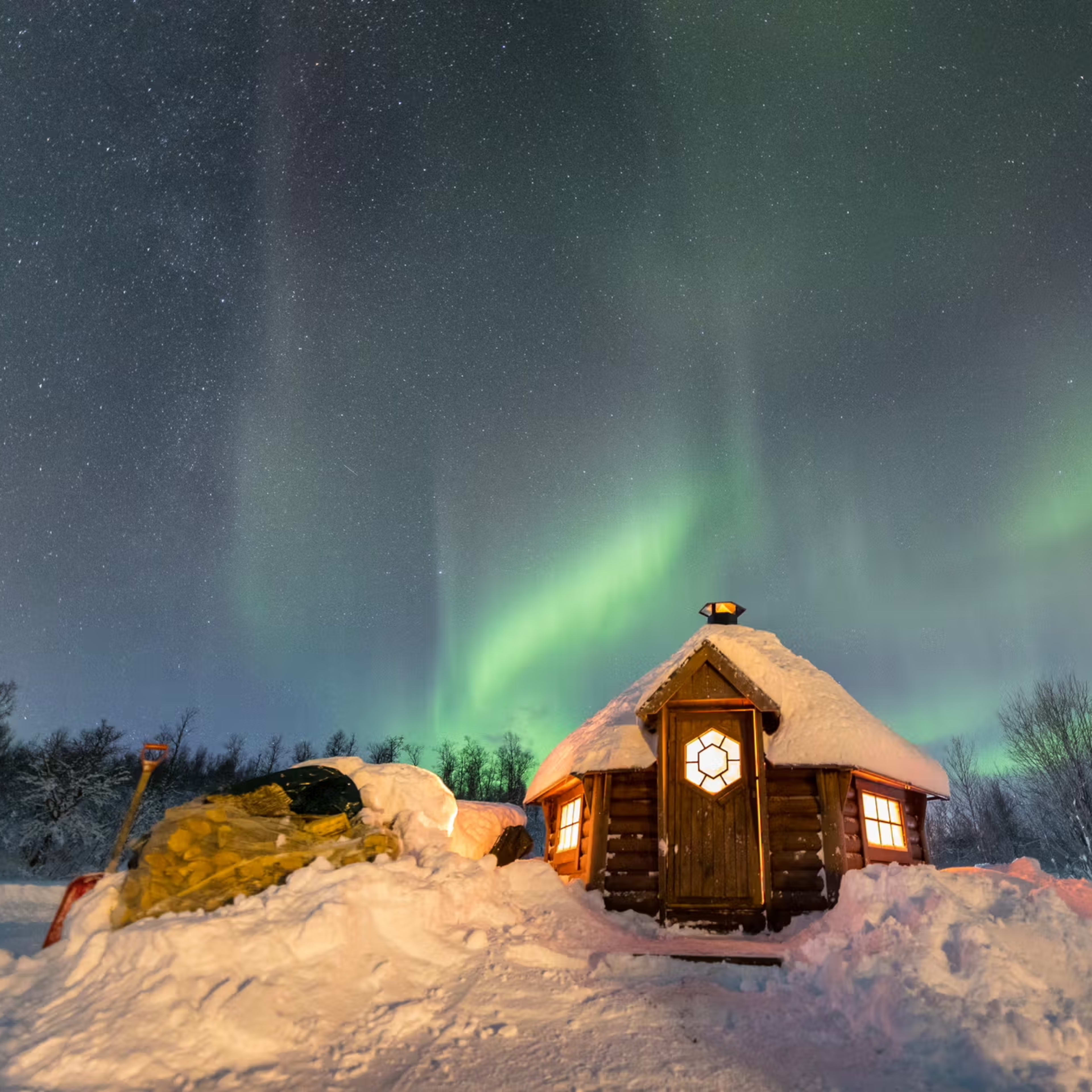 Varm trehytte lyser i snølandskap med grønne nordlys over