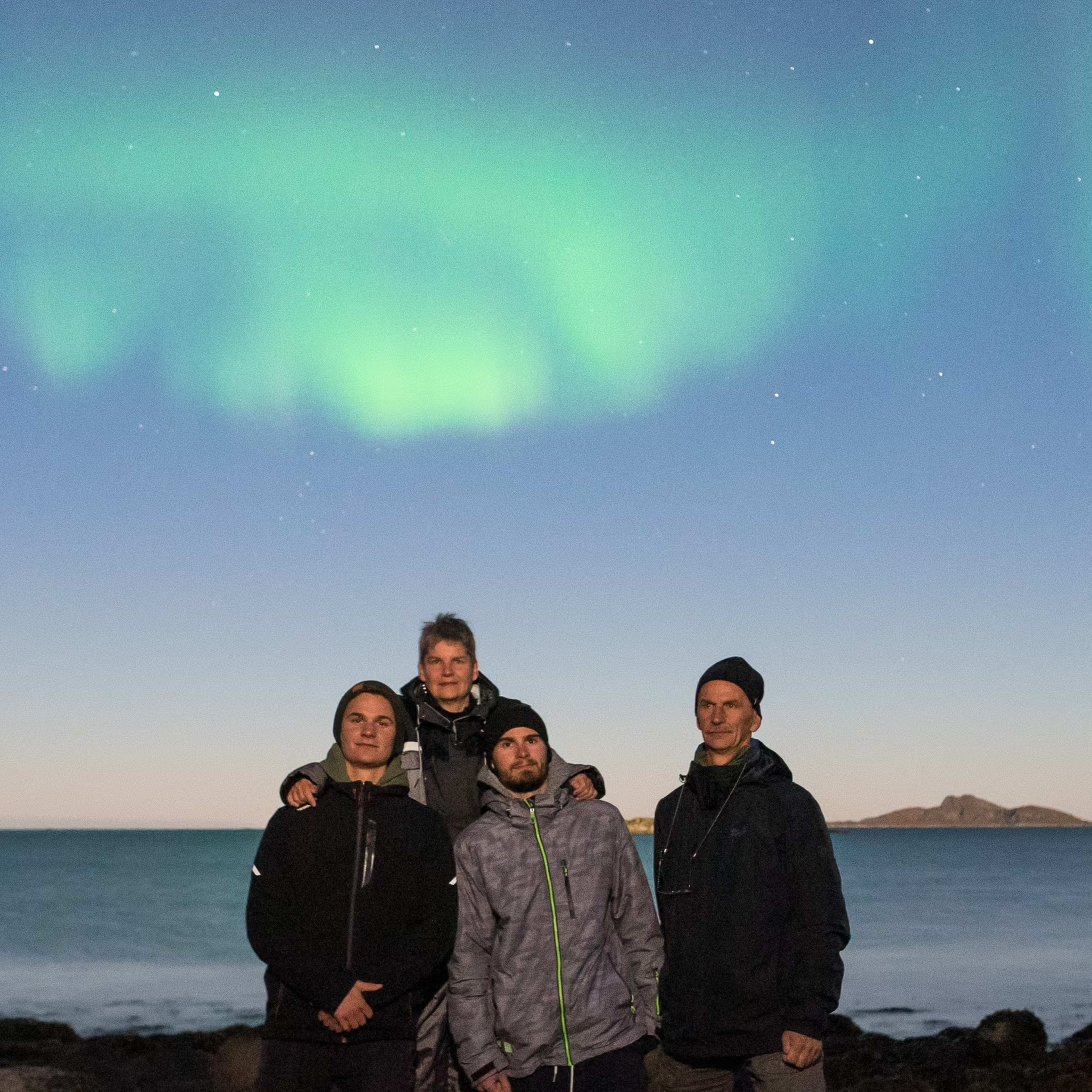 Fire familiemedlemmer på steinete kyst med grønne nordlys som speiler seg i vannet
