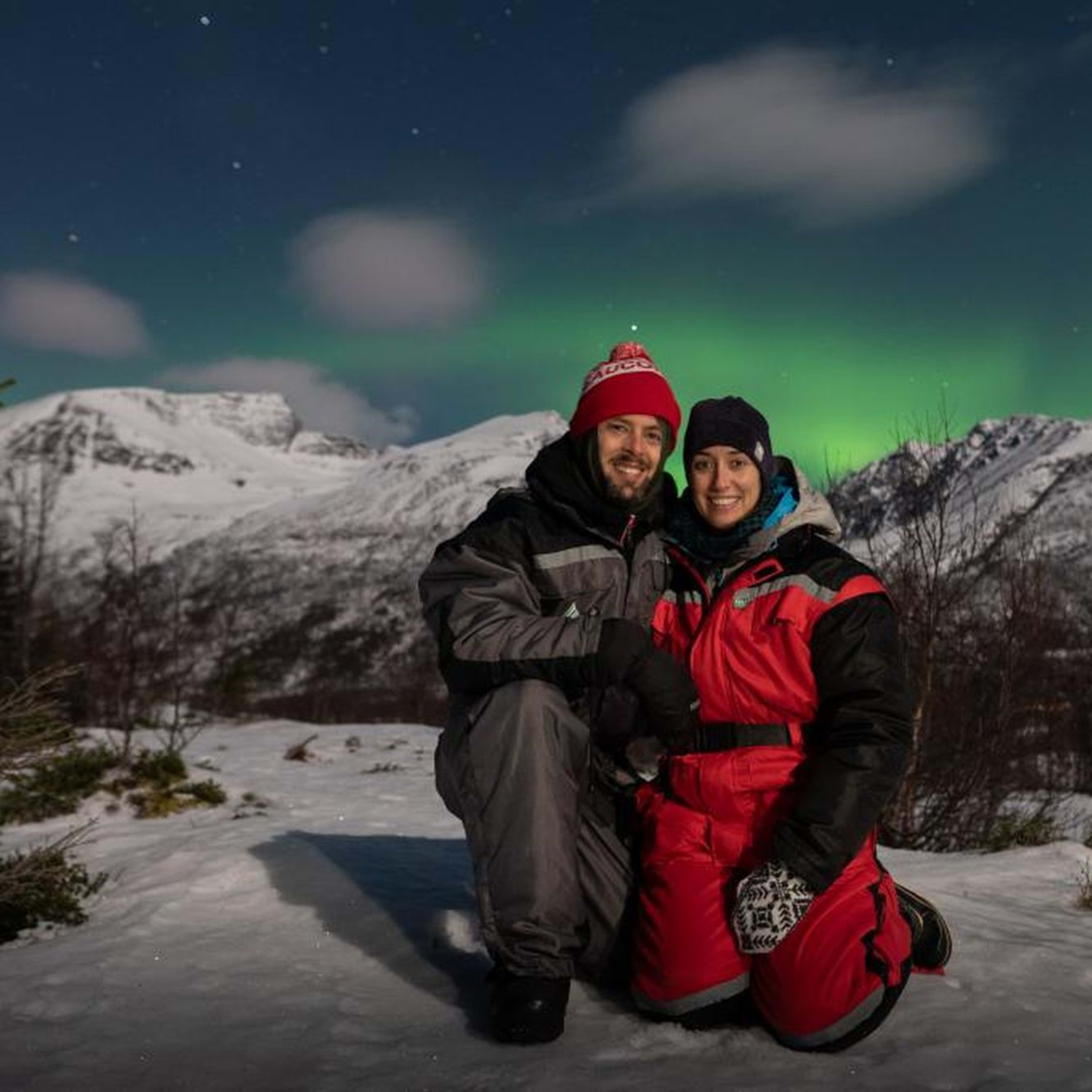 Ein lächelndes Paar kniet im Schnee unter dem Nordlicht, mit einer verschneiten Berglandschaft im Hintergrund.