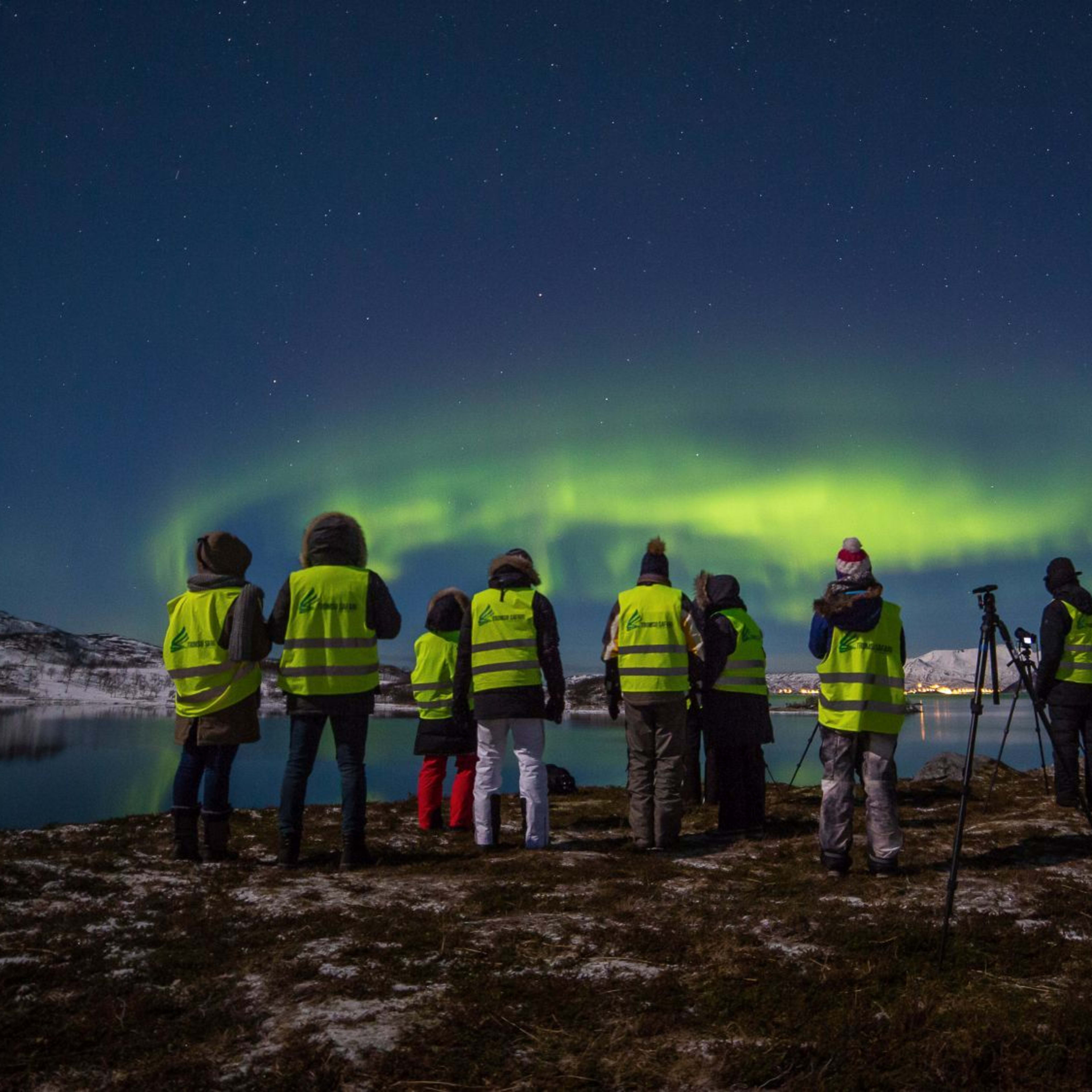 Folk i gule vester ser på nordlyset over et snødekt landskap med innsjø og fjell i bakgrunnen.
