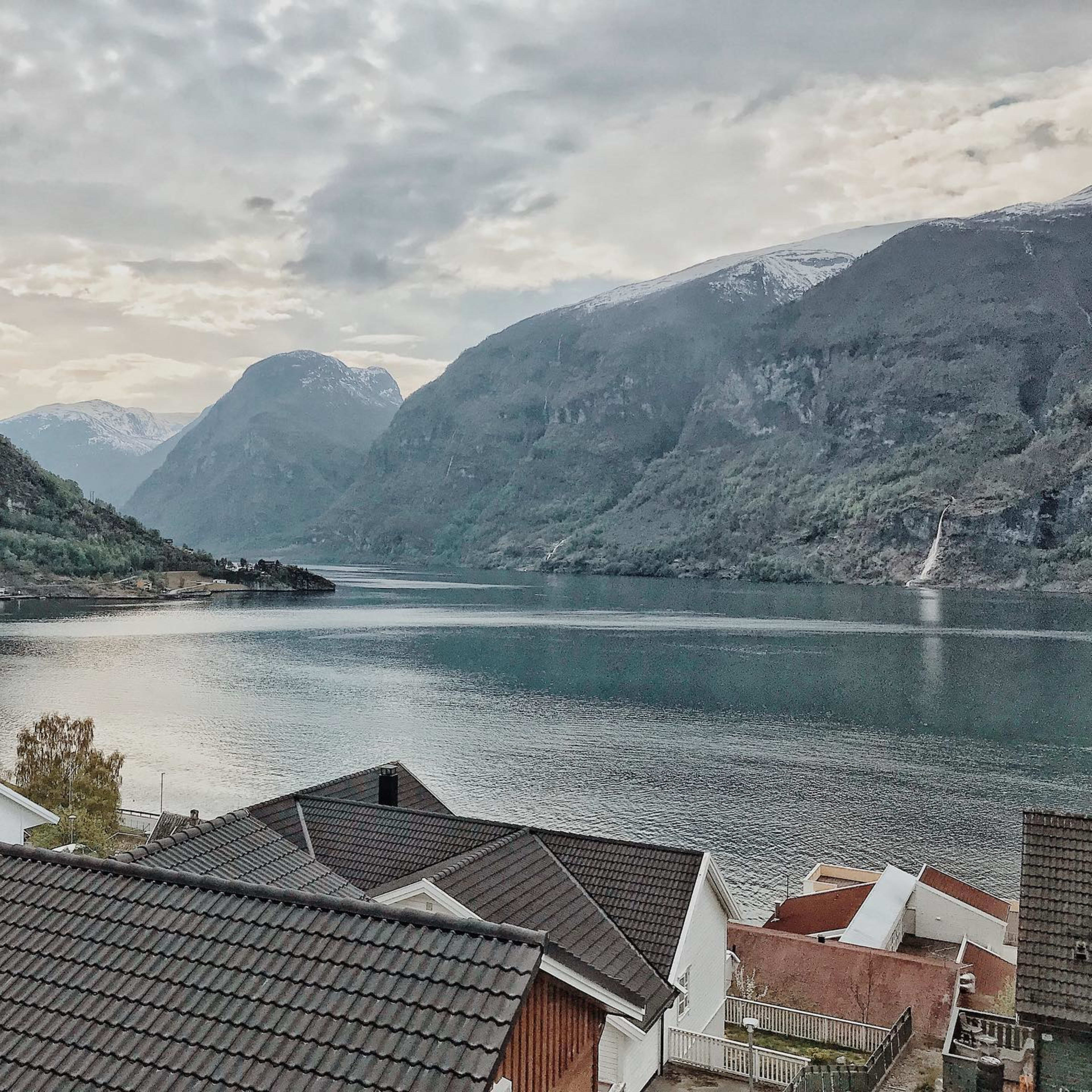 Ruhiger Fjord mit Bergen und Häusern bei bewölktem Himmel.