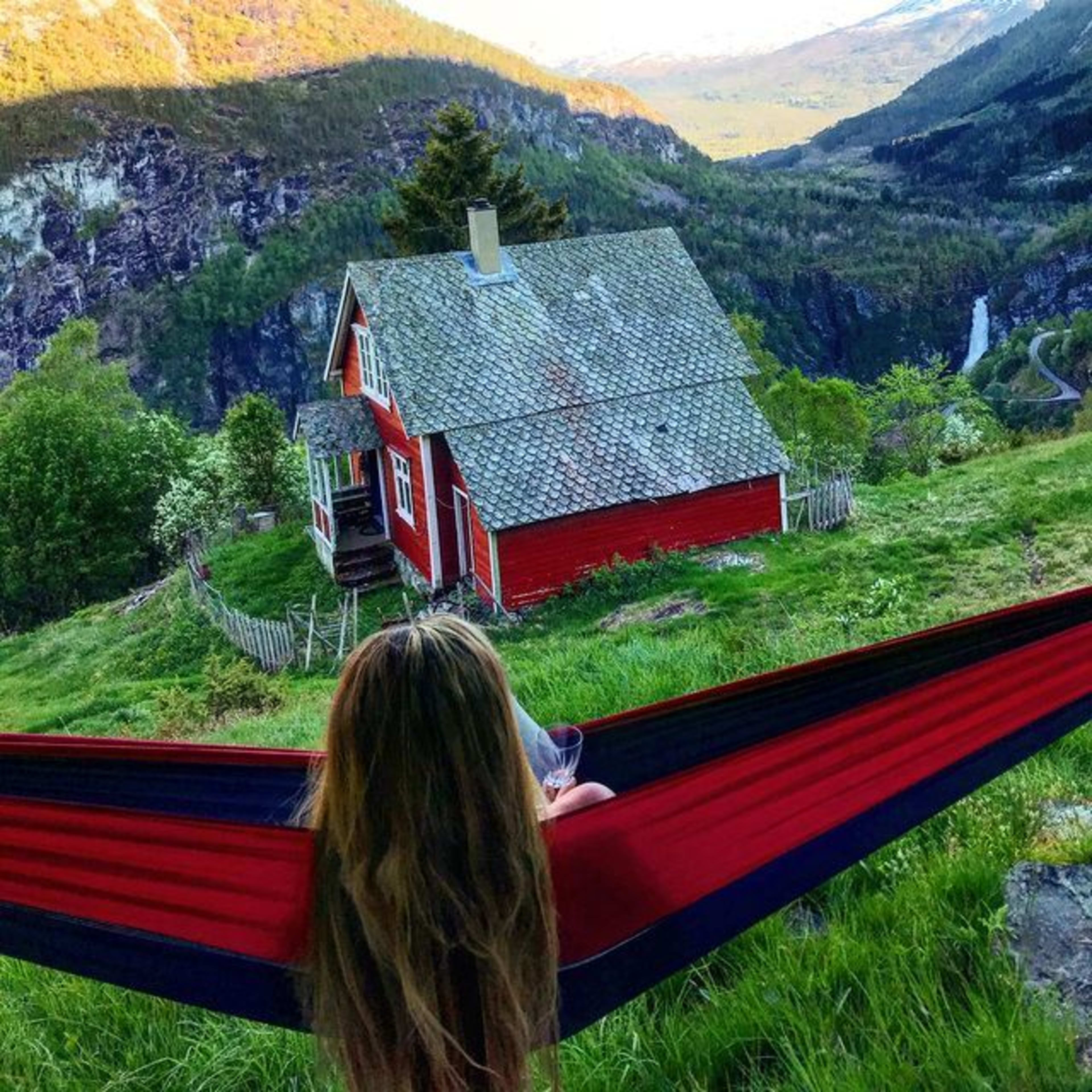 Woman in hammock looks at red cottage in golden mountain light.