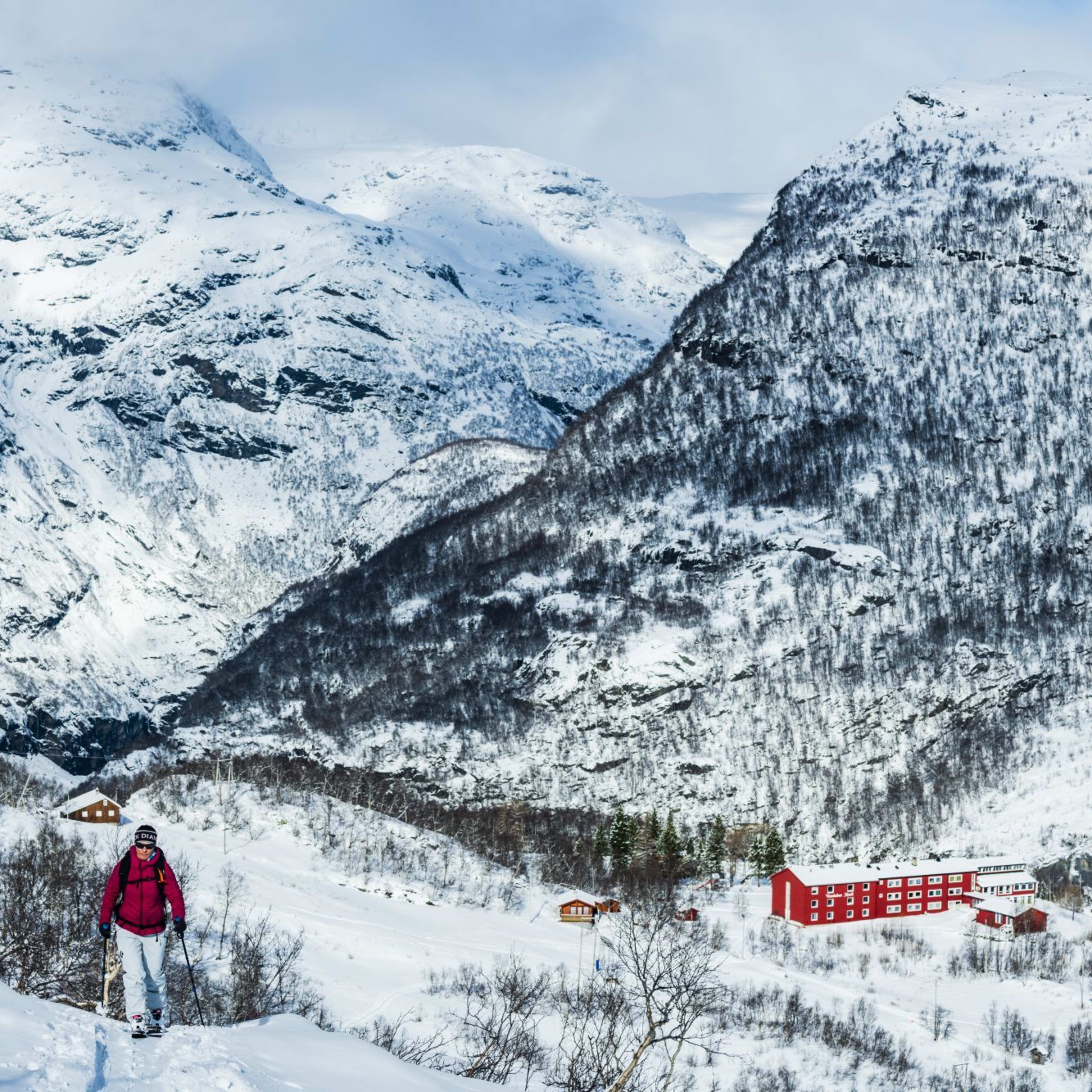 Skiløper i rød jakke går i snø, omgitt av røde hytter og fjell.