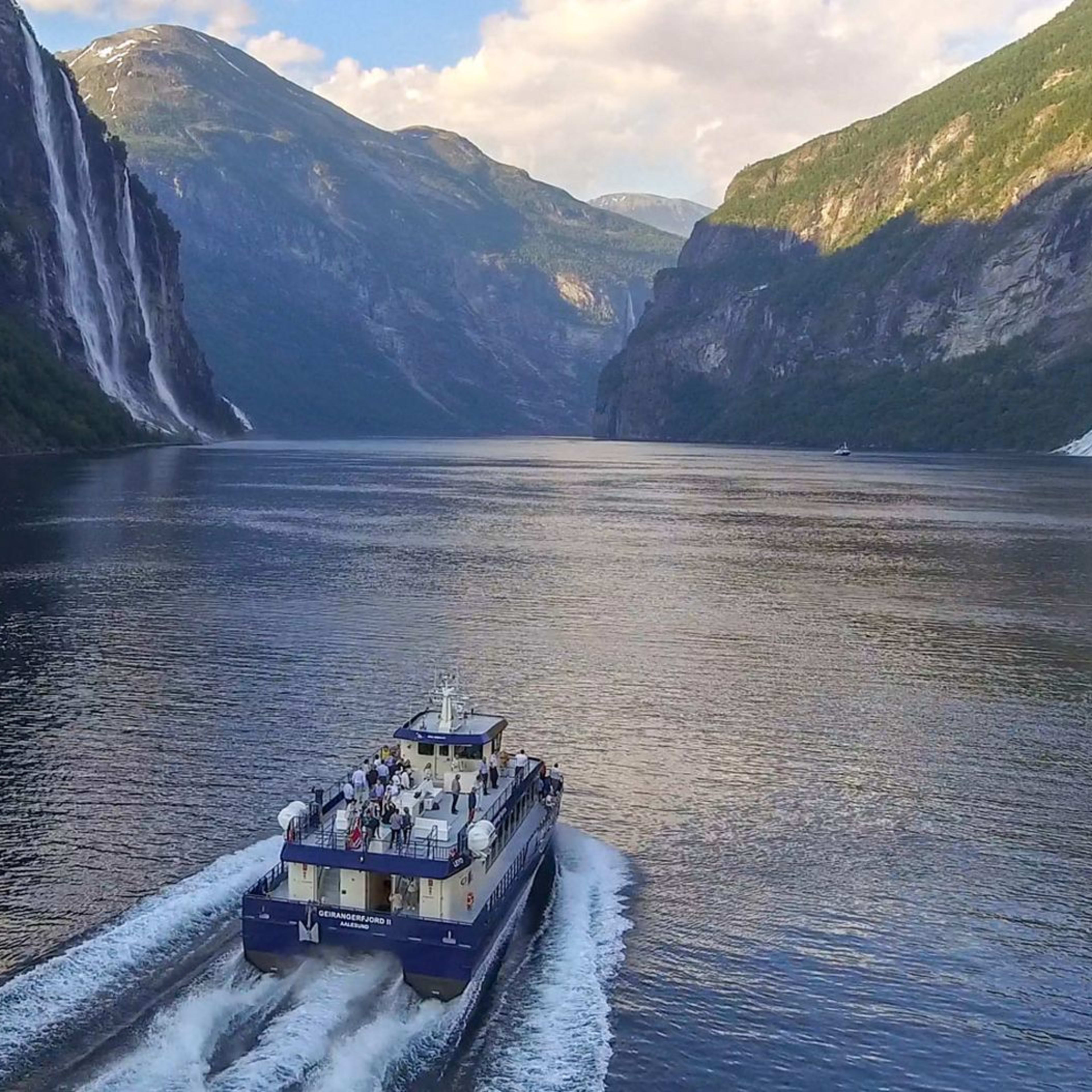 Boot fährt durch engen Fjord mit üppigen Klippen, Wasserfällen und ruhigem, blauem Wasser, das die Landschaft spiegelt.