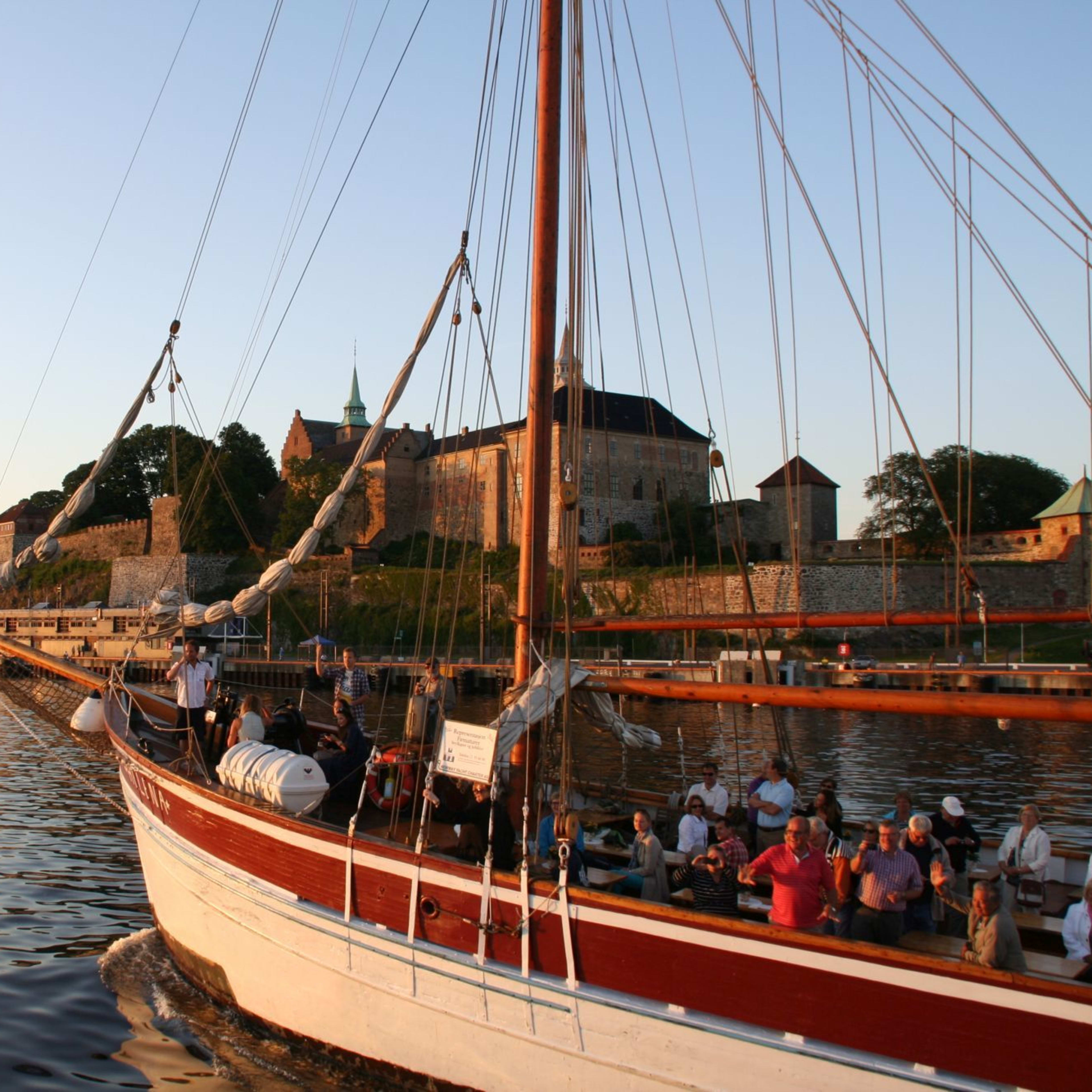 Turister på fjordcruise på Oslofjorden passerer historiske Akershus festning ved solnedgang.
