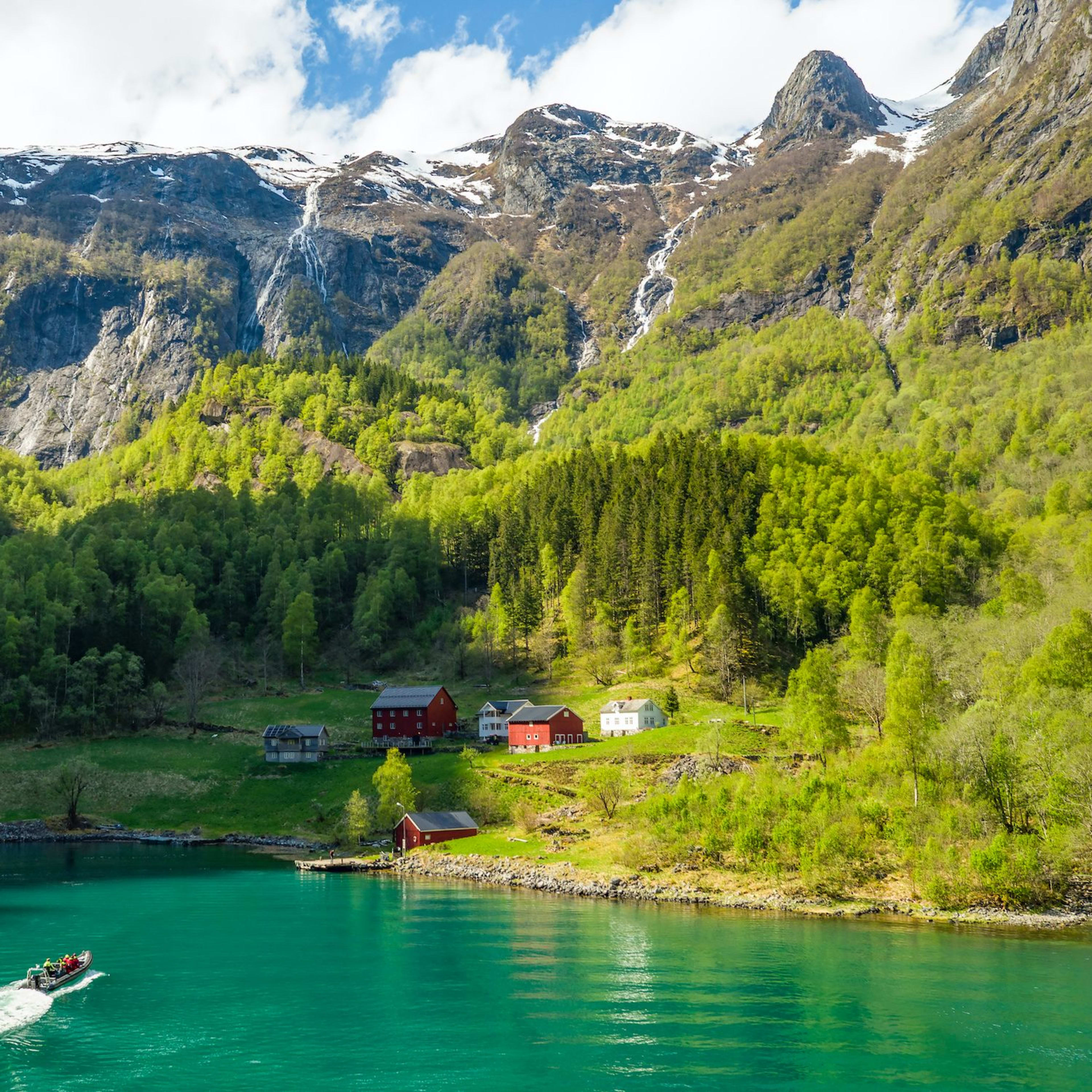 Motorboat on turquoise fjord heading toward village with houses nestled against forested hillside.