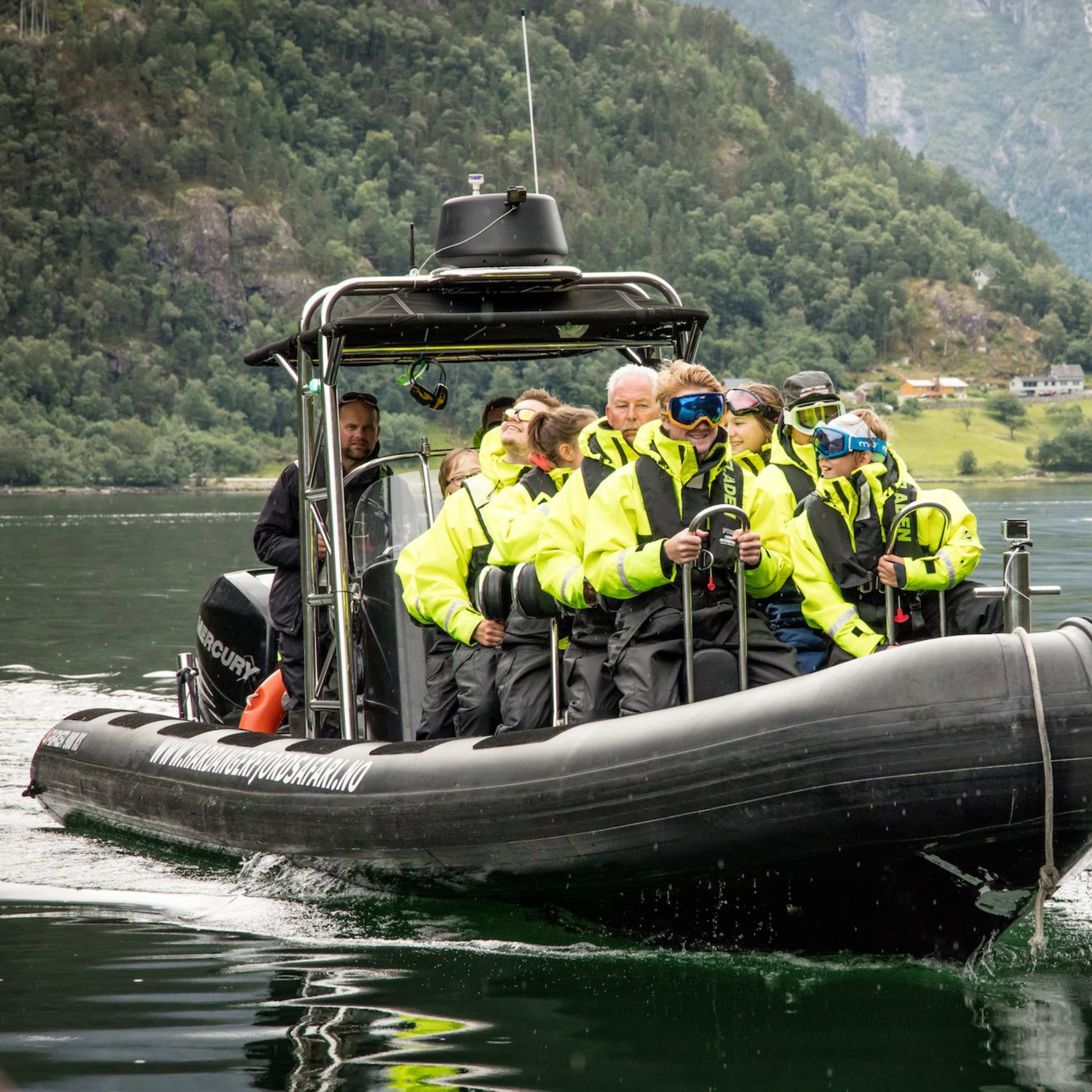 Tourists in yellow ride a boat through fjord with green hills and village.