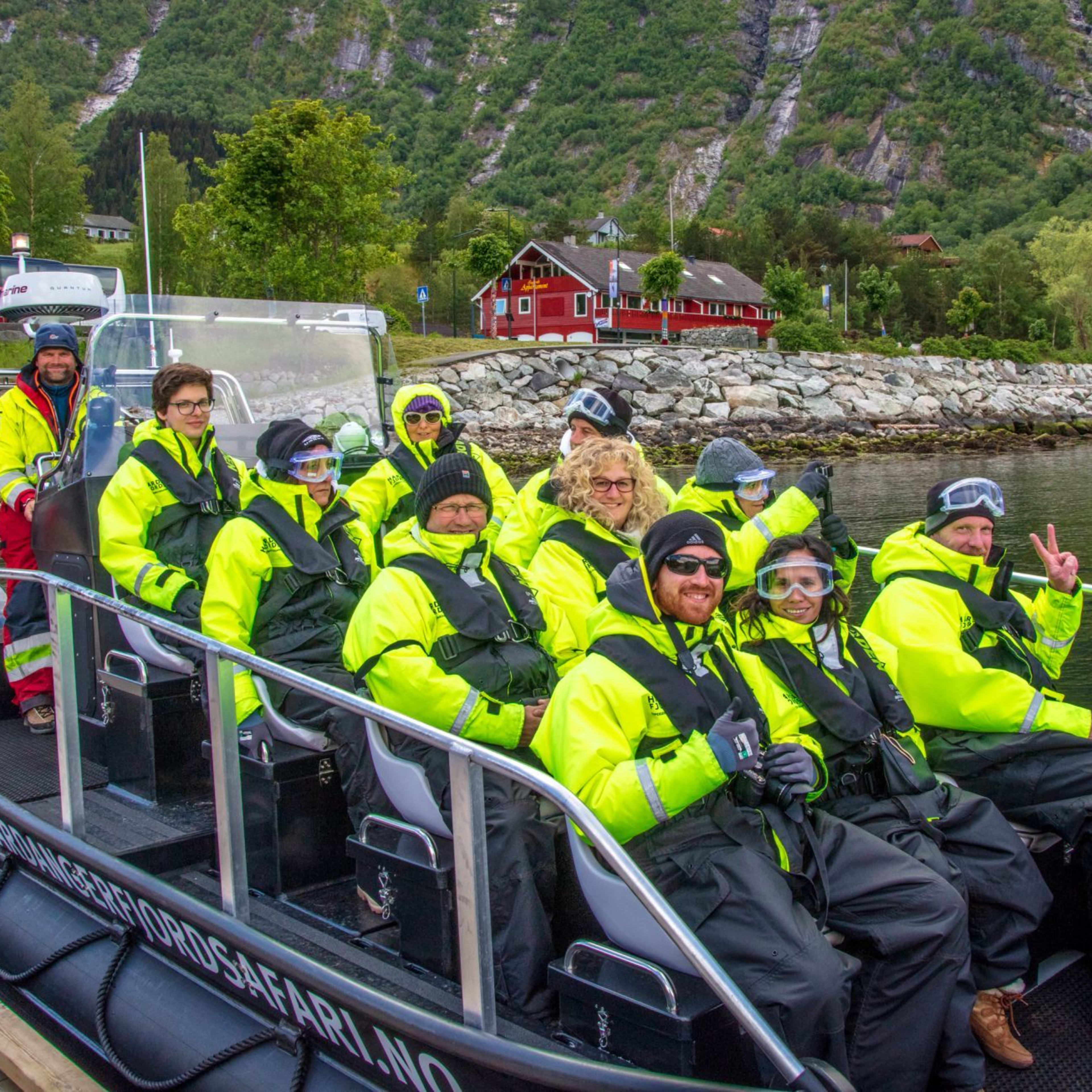 Smiling tourists in yellow suits sit in a black boat near shore and red cabin.