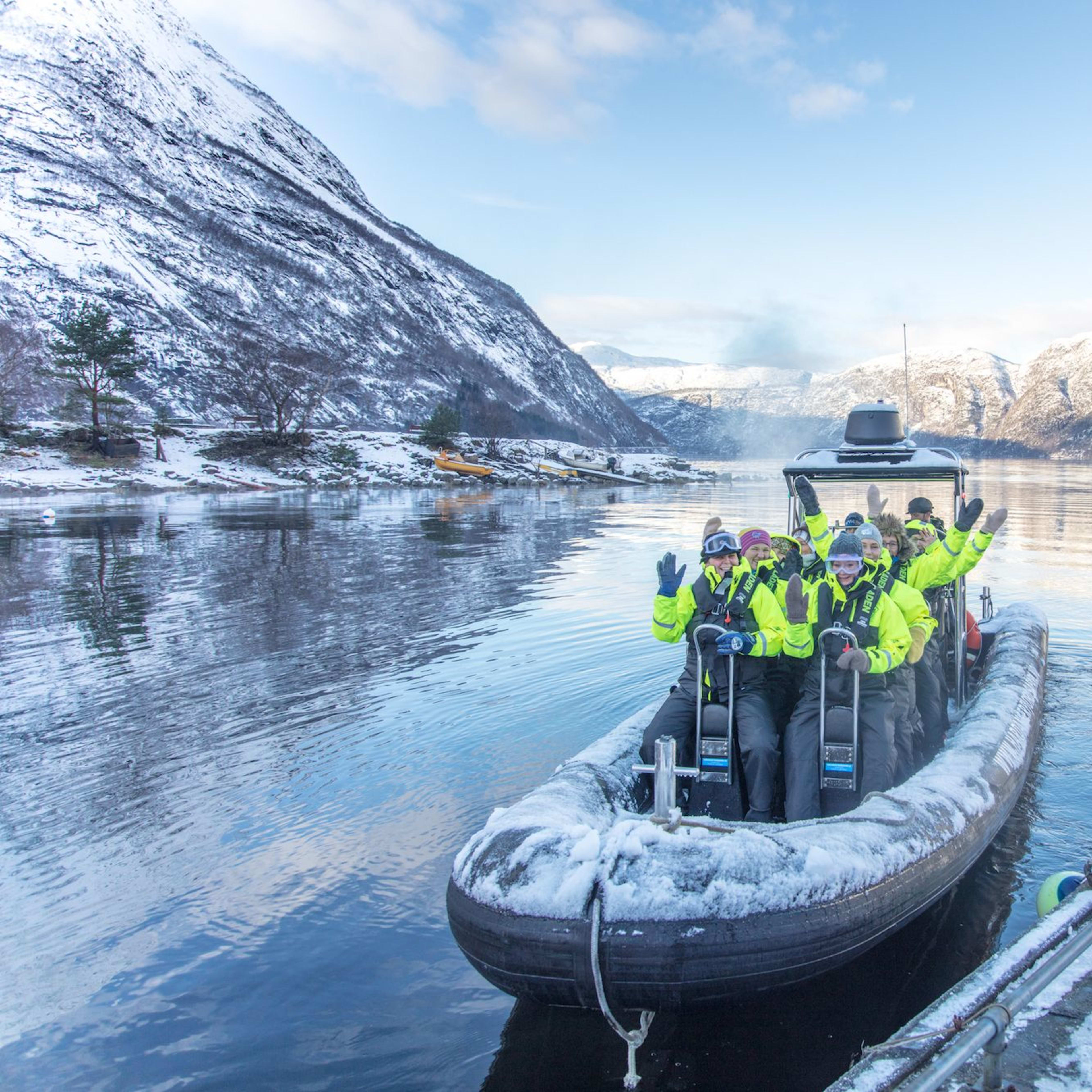 People wave from snow-covered RIB boat on calm winter fjord.
