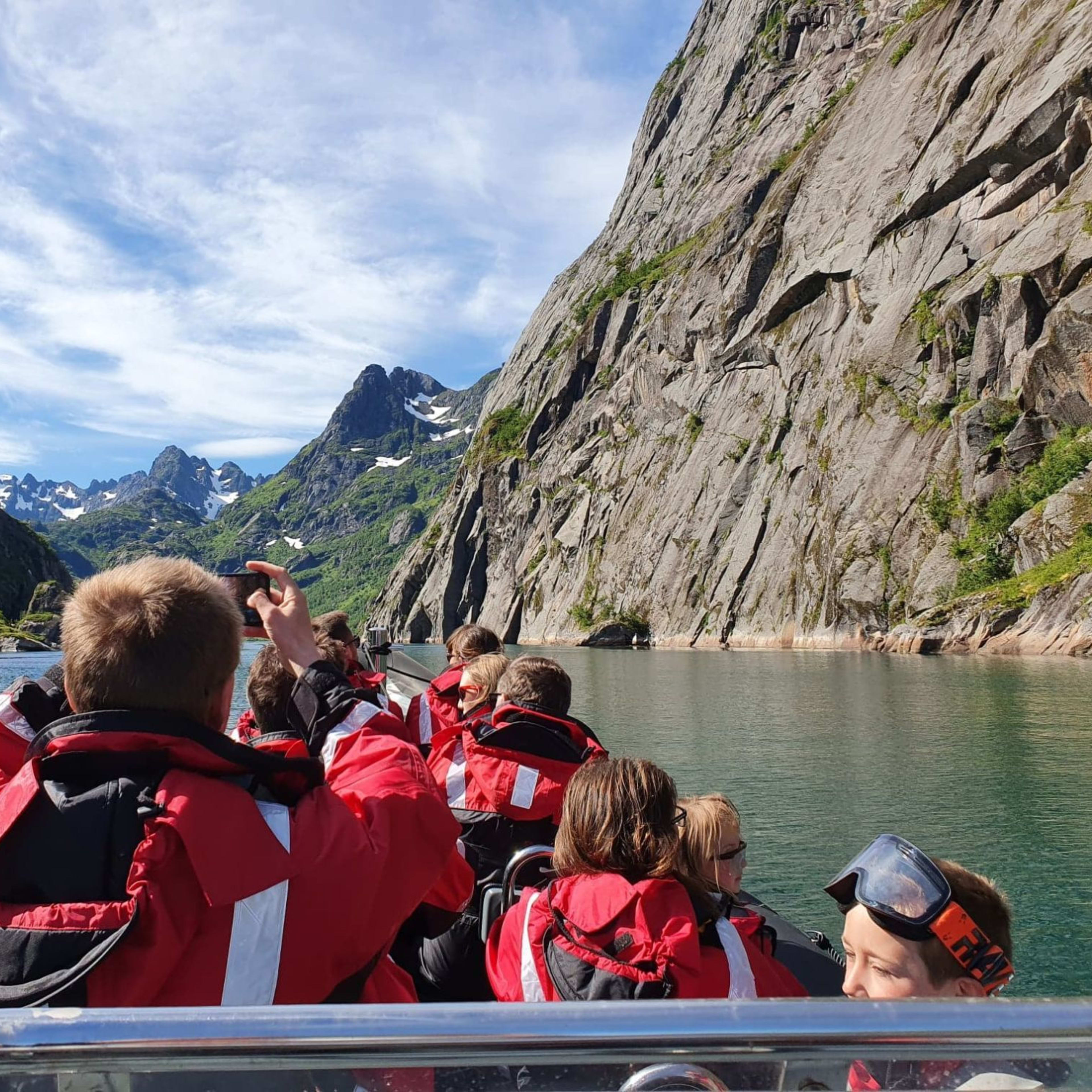 Group in red and black waterproof suits on RIB cruising through calm fjord surrounded by cliffs and snow-capped peaks.