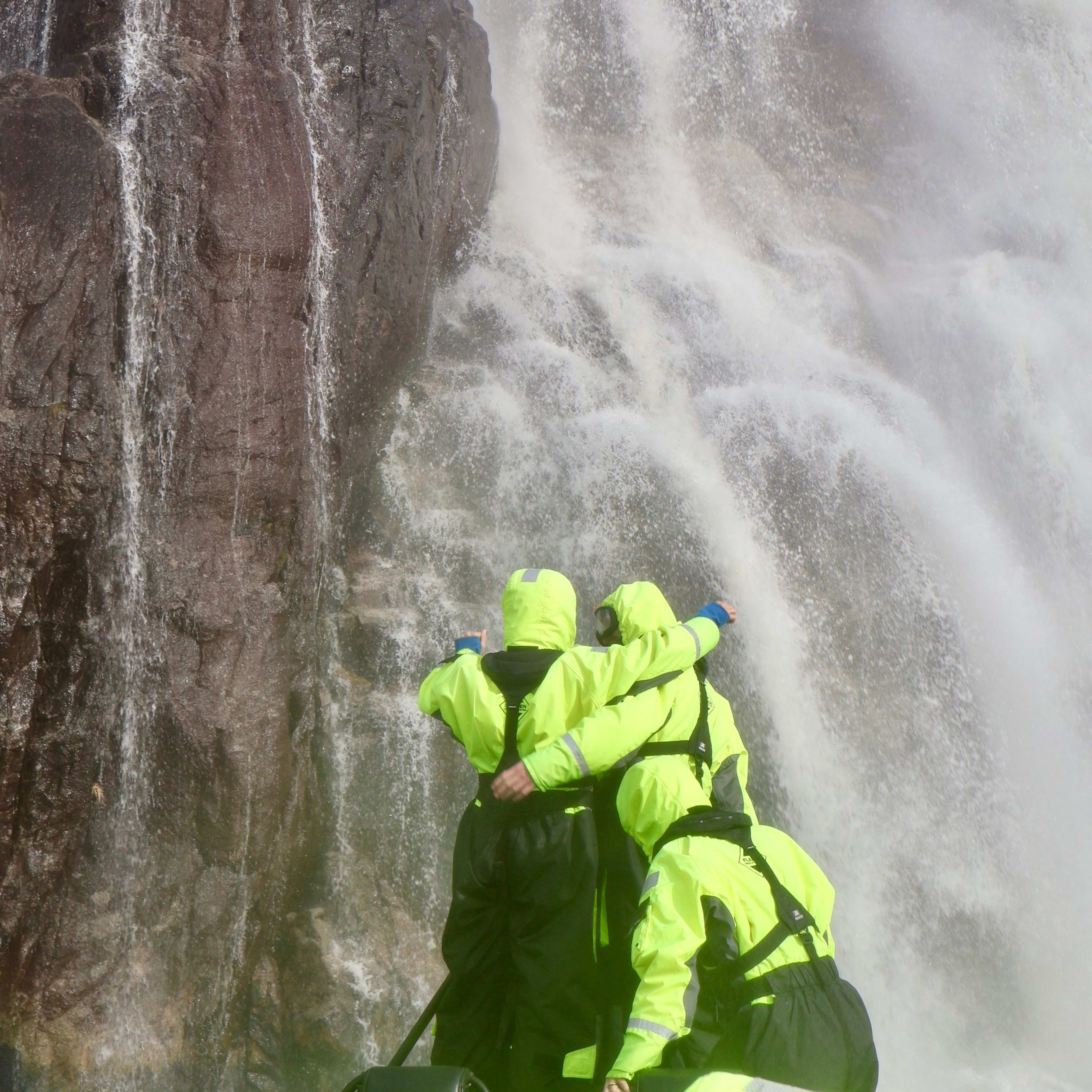 Drei Passagiere in leuchtend neon-grünen Jacken stehen auf einem Schnellboot und strecken sich zu einem mächtigen Wasserfall.